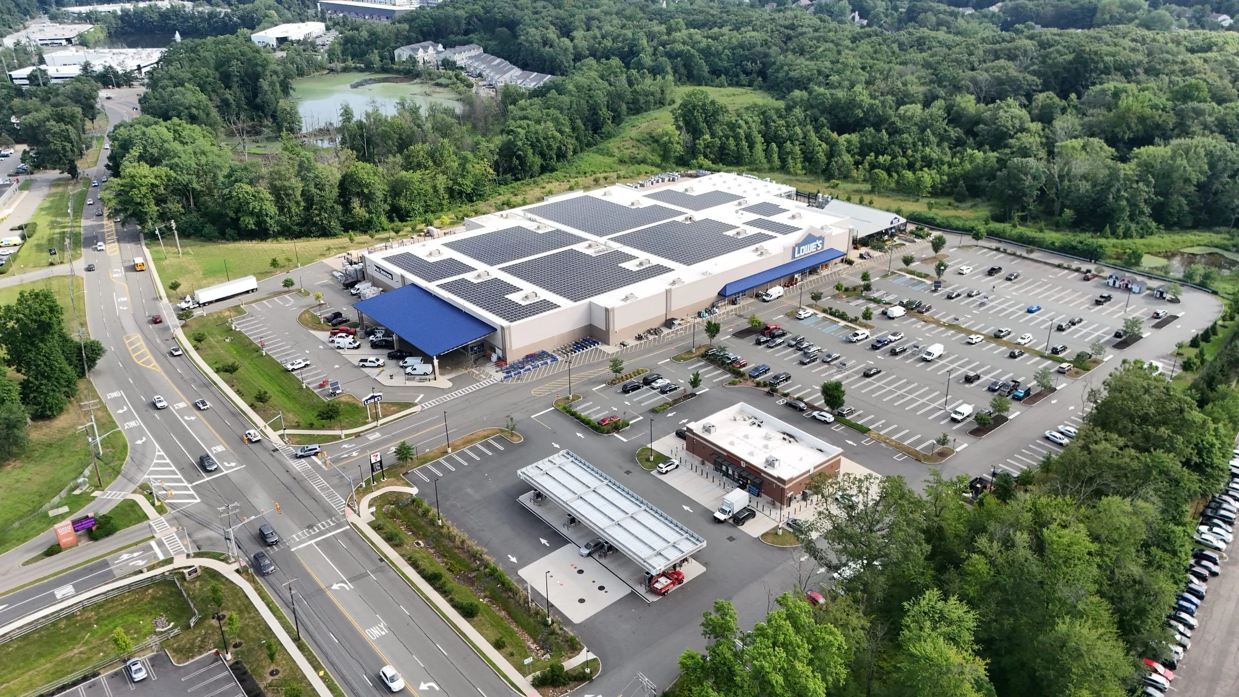 Aerial view of a large Lowe's hardware store with solar panels on the roof, surrounded by a parking lot filled with cars, adjacent to roads, trees, and green areas.