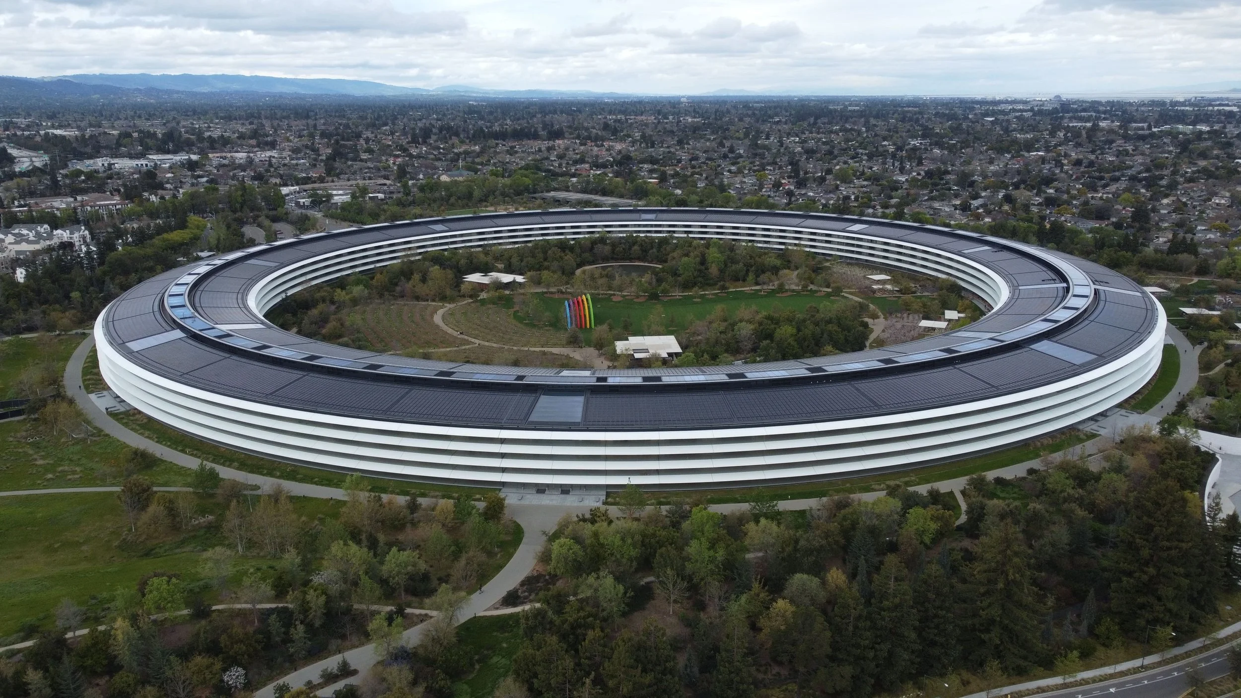 Aerial view of the Apple Park headquarters in Sunnyvale, California, featuring its distinctive circular building with solar panels on the roof and surrounded by green landscape and trees.