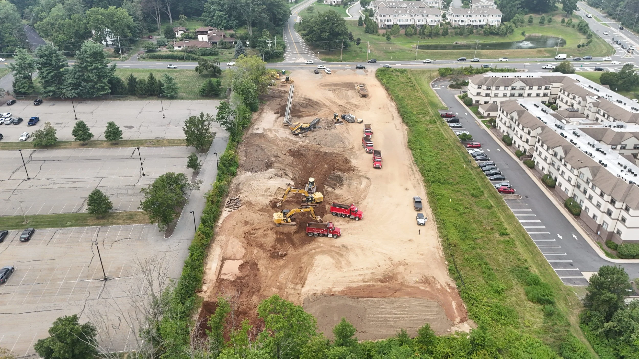 Construction site with excavators and dump trucks, partially cleared land, surrounded by parking lots, residential buildings, and greenery.