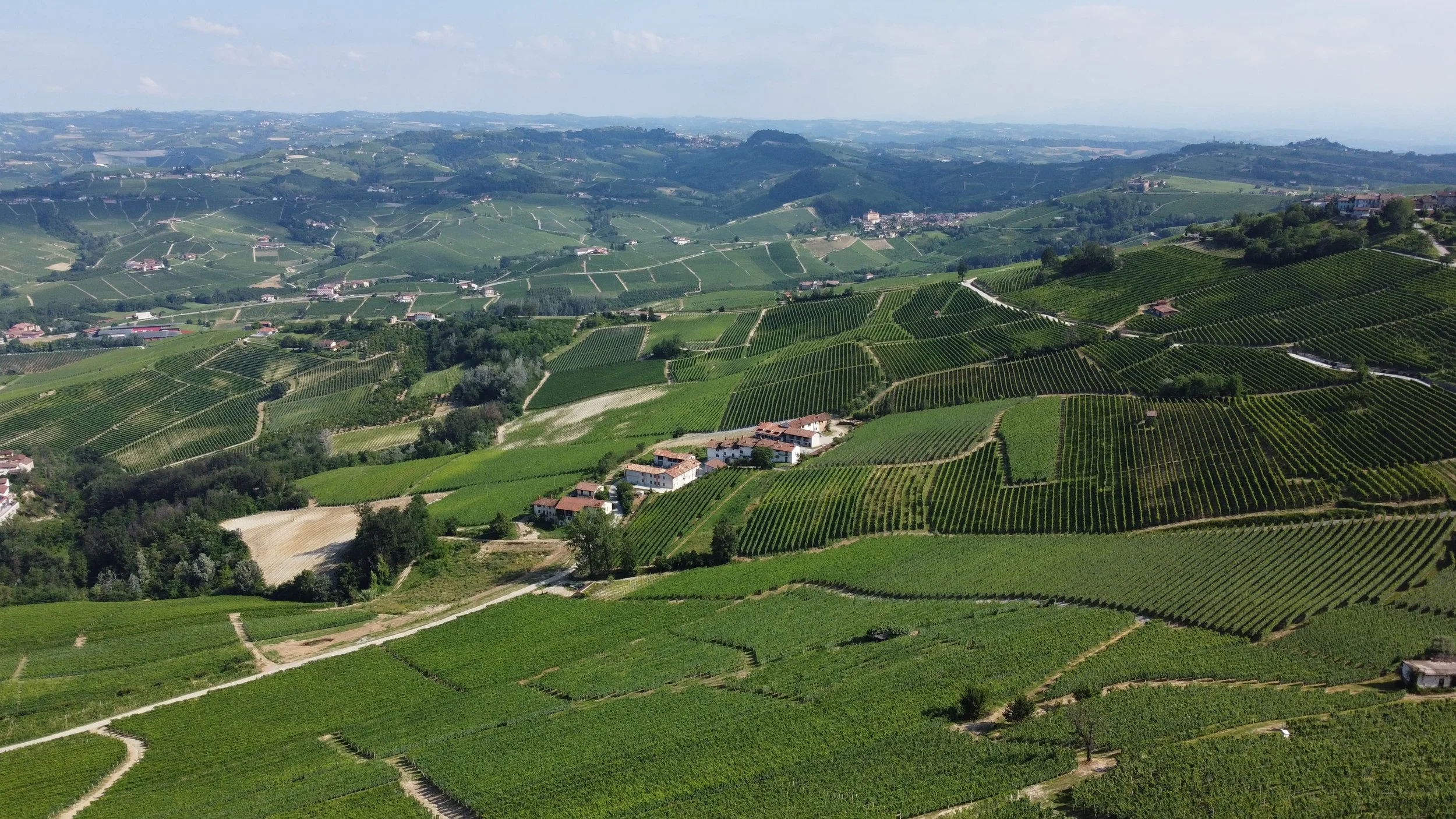 Aerial view of green rolling hills and vineyards with small farm buildings, winding dirt roads, and distant hills under a partly cloudy sky.