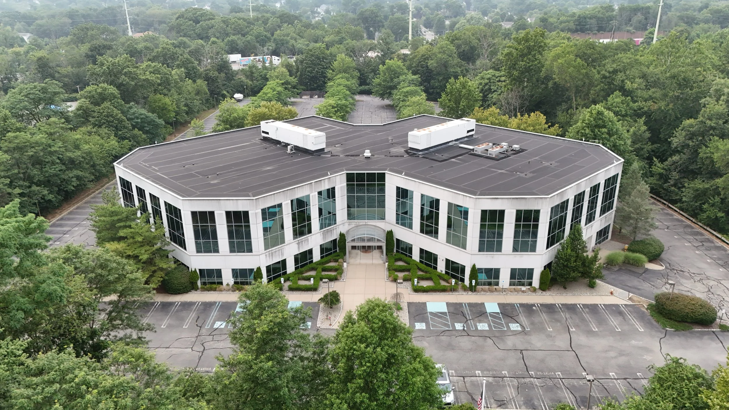 An aerial view of a modern, two-story office building with large glass windows, a landscaped entrance, and an empty parking lot surrounding it. The building has a curved shape and a black roof with HVAC units.