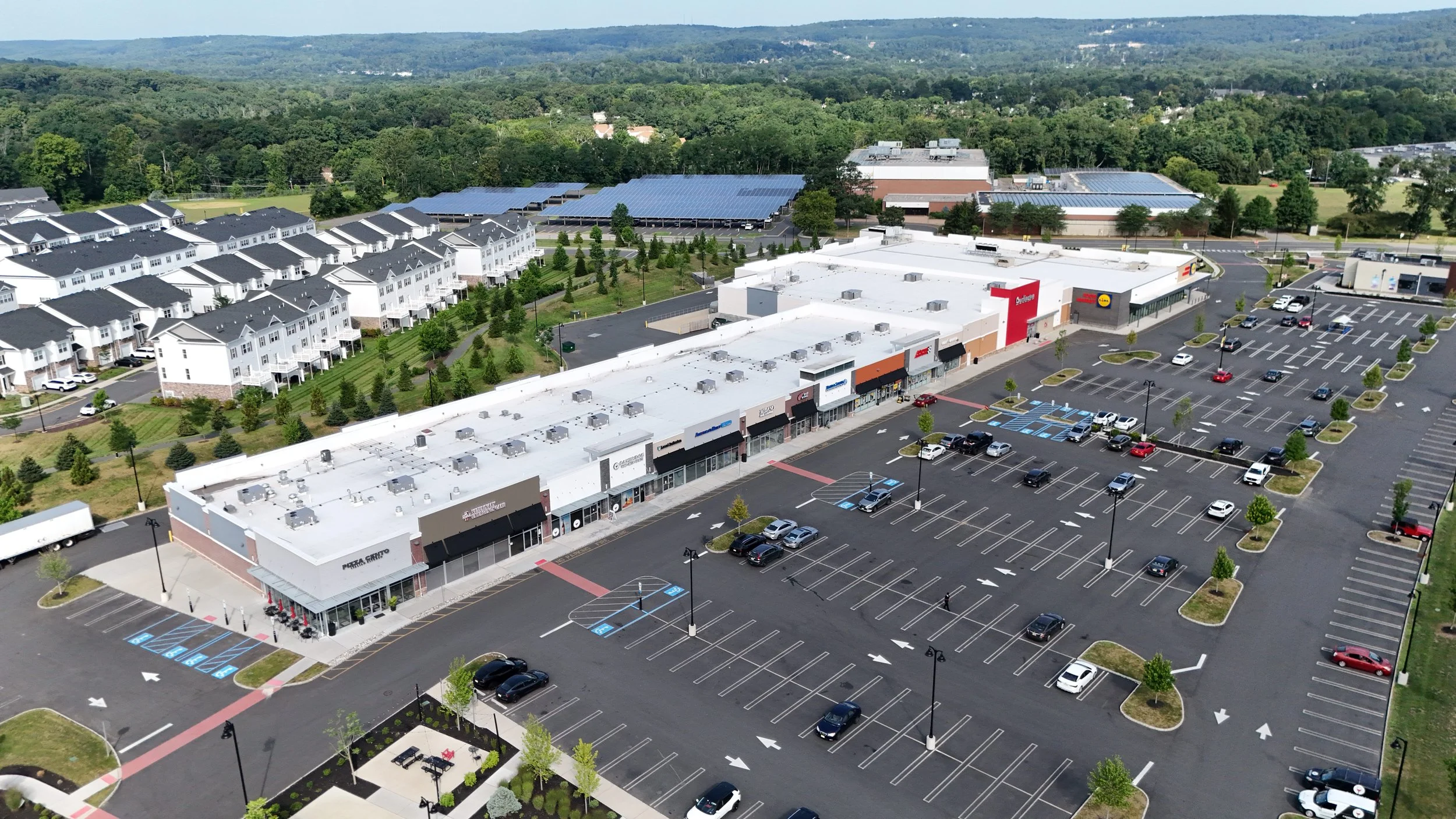 Aerial view of a strip mall with various retail stores, surrounded by a large parking lot with some cars, empty spaces, and several trees, with a residential area and green landscape in the background.