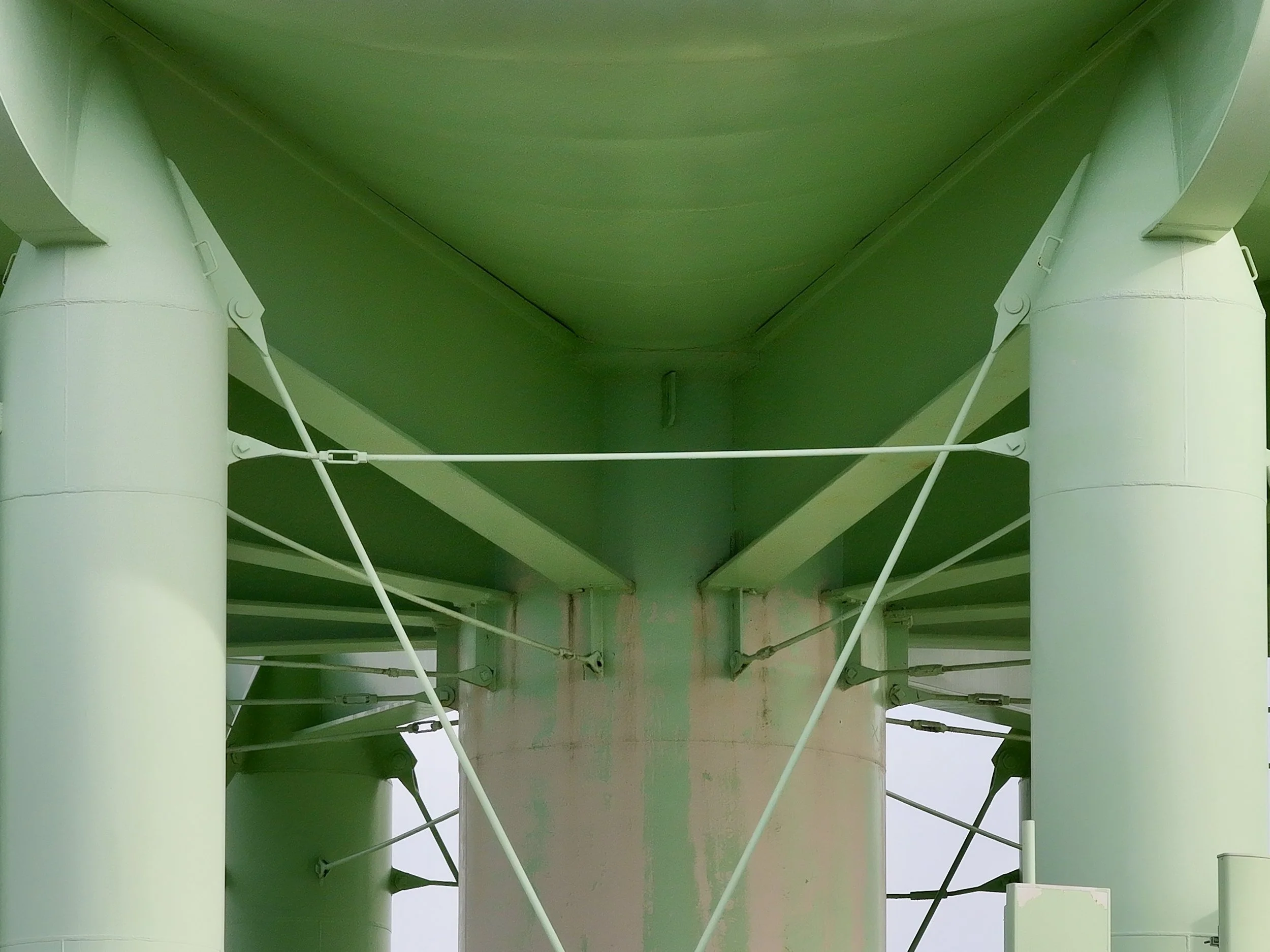 Underneath a large green bridge with multiple white support beams, view from below showing the underside of the structure with pipes and cables.