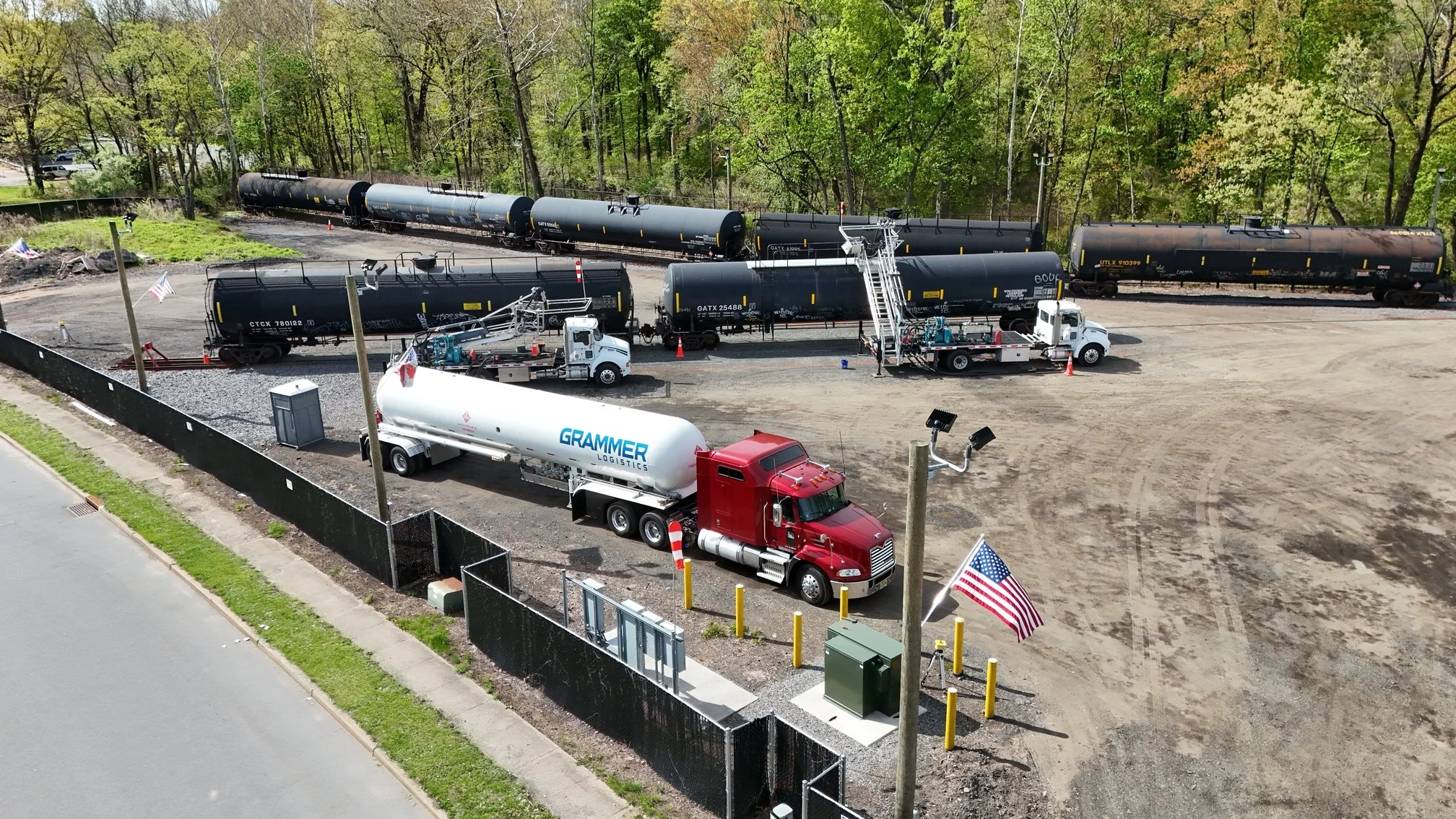 Aerial view of a train yard with black tanker cars and two trucks, one white with a logo and one red, parked on a gravel lot near a chain-link fence, with trees and a road in the background.
