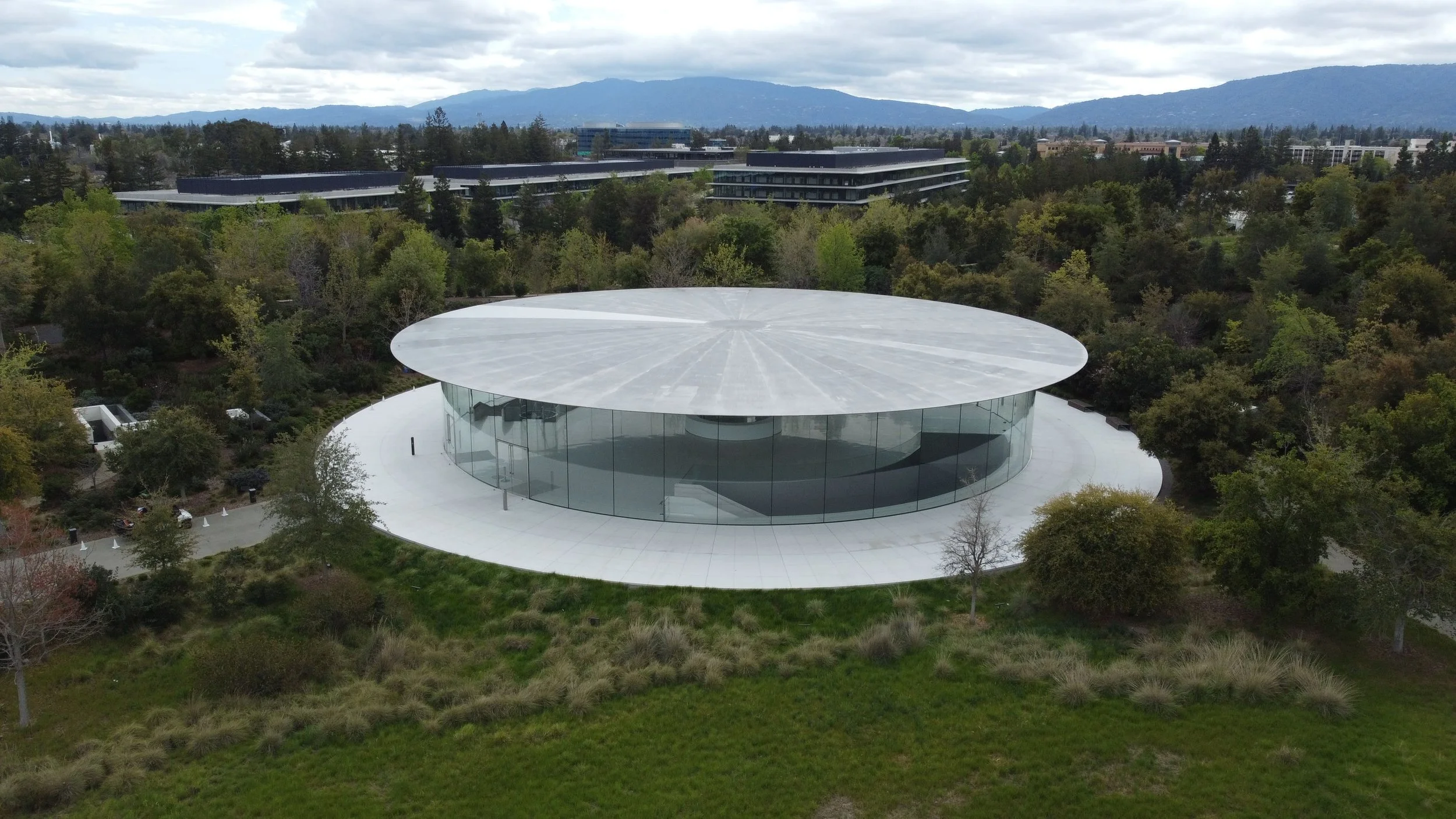 An aerial view of a circular modern glass building with a flat, circular roof, surrounded by greenery and trees, with mountains and additional modern buildings in the background.