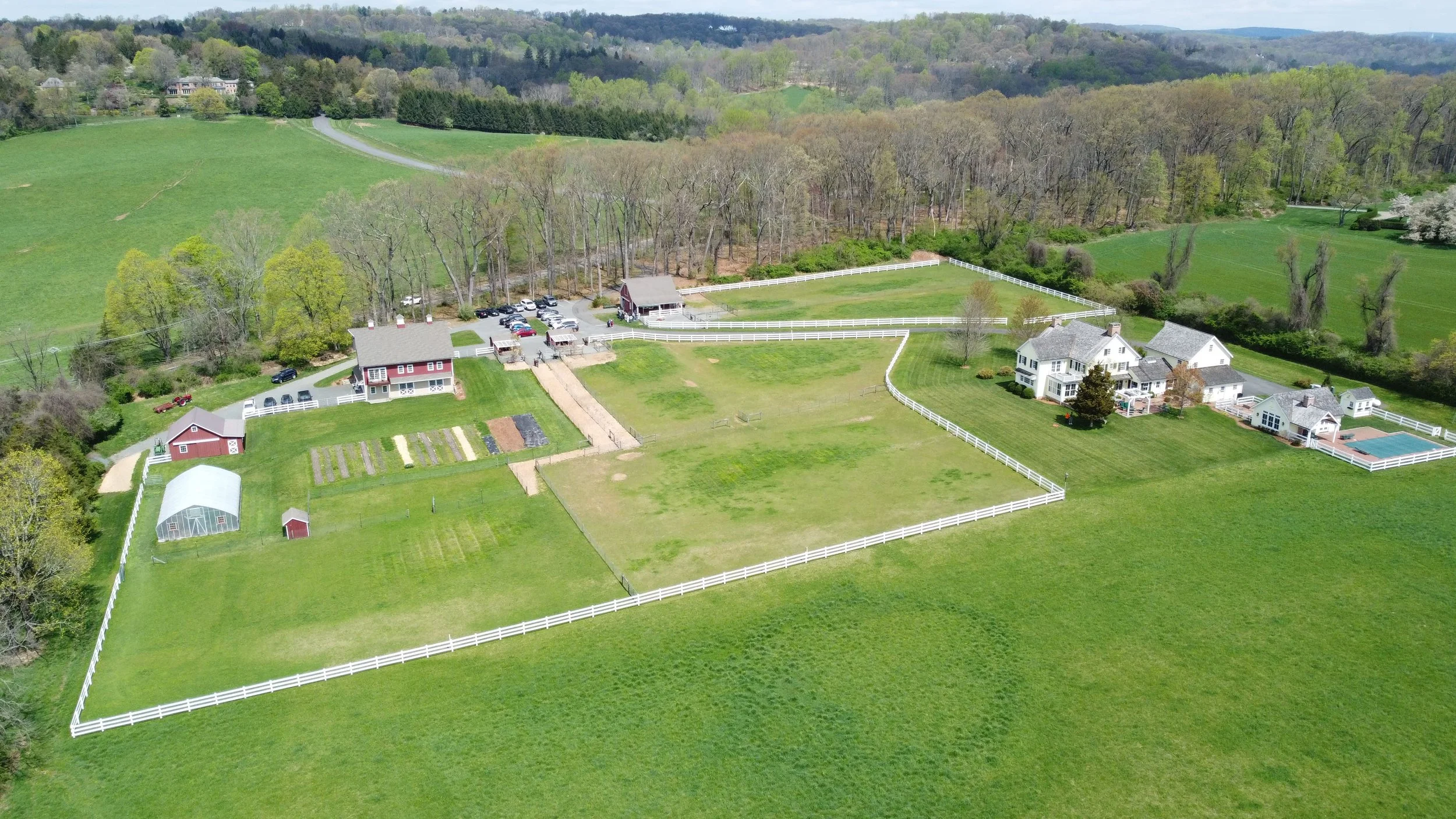 Aerial view of a large countryside property featuring a white house, a guest house, a fenced yard, a garden with planting beds, a greenhouse, and several outbuildings, all surrounded by green fields and trees.