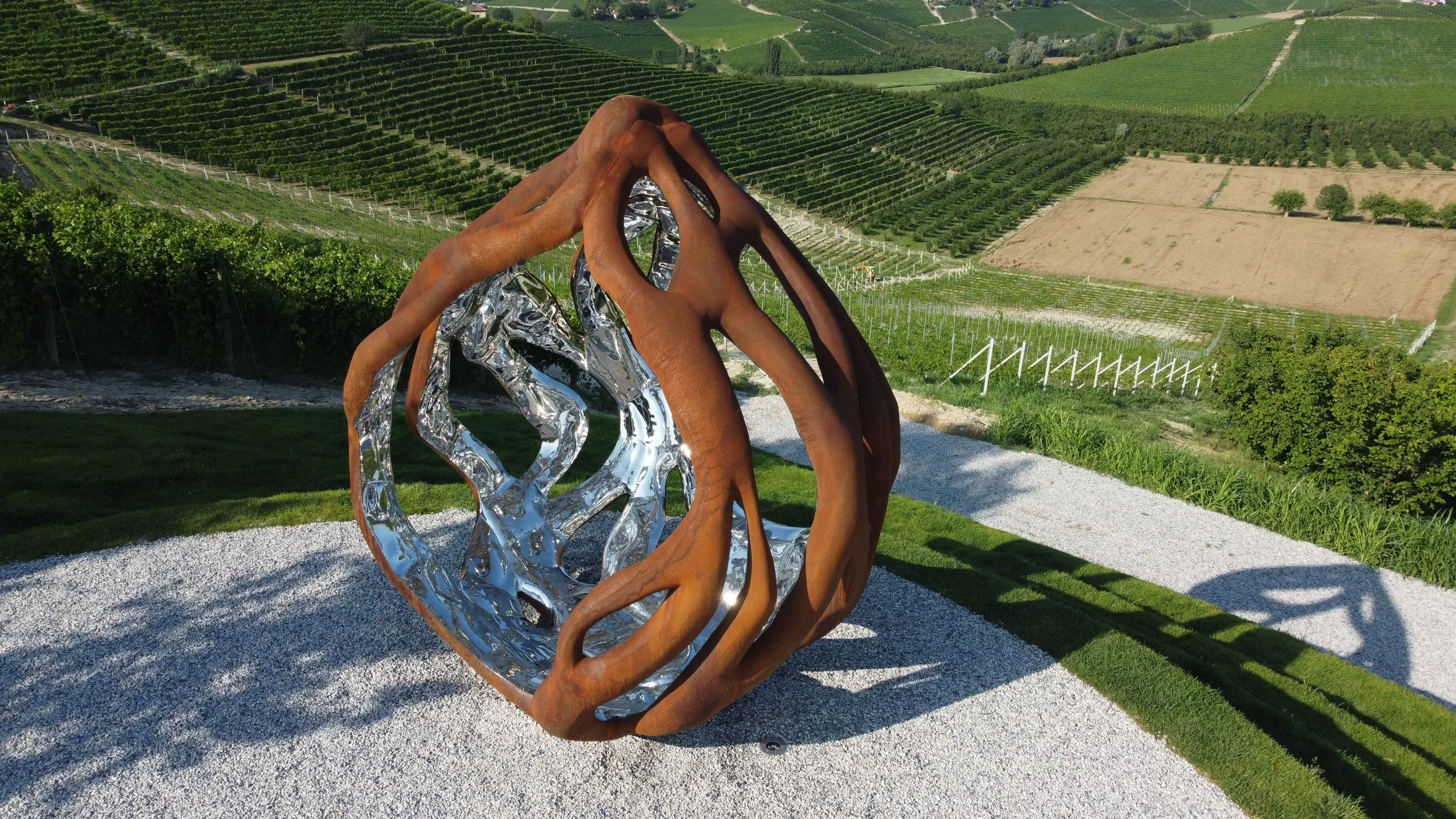 Outdoor sculpture with a wooden and metallic structure on a gravel surface, overlooking a lush green hillside with vineyard rows.