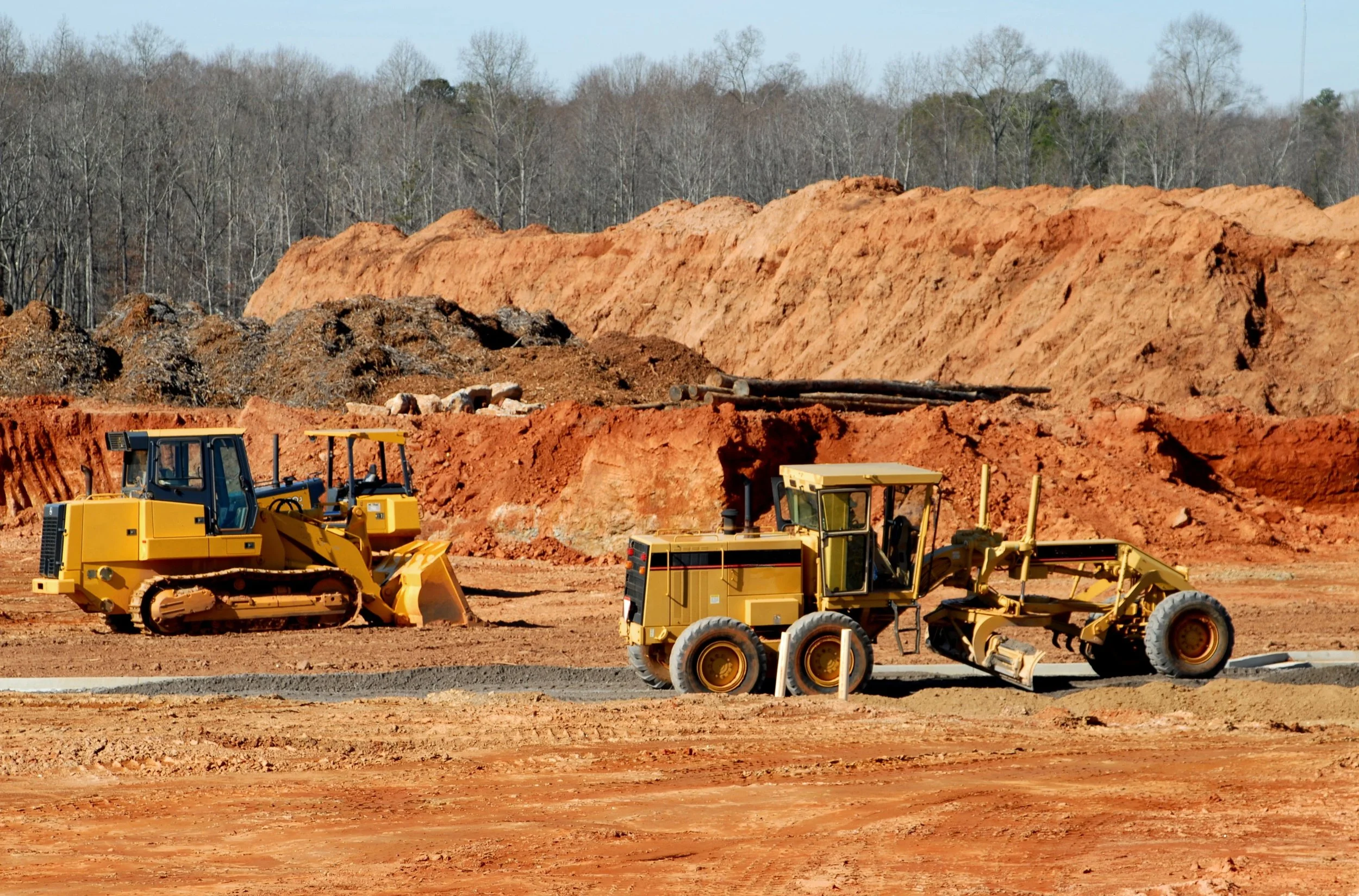 Construction site with two yellow bulldozers working on reddish dirt and sand, with trees and clear sky in the background.