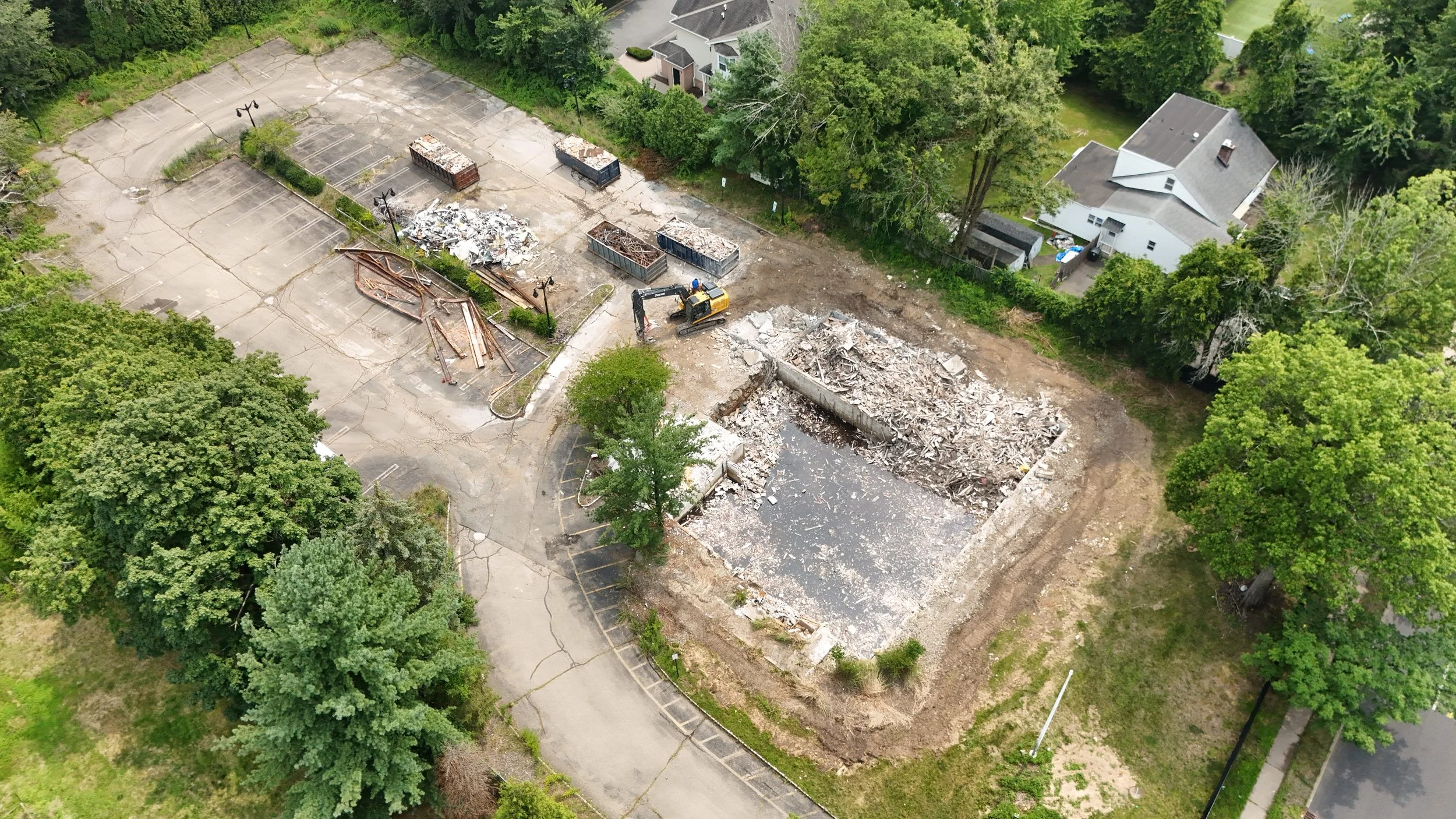Aerial view of a construction or demolition site with an excavator, rubble, and debris, neighboring residential houses and greenery.
