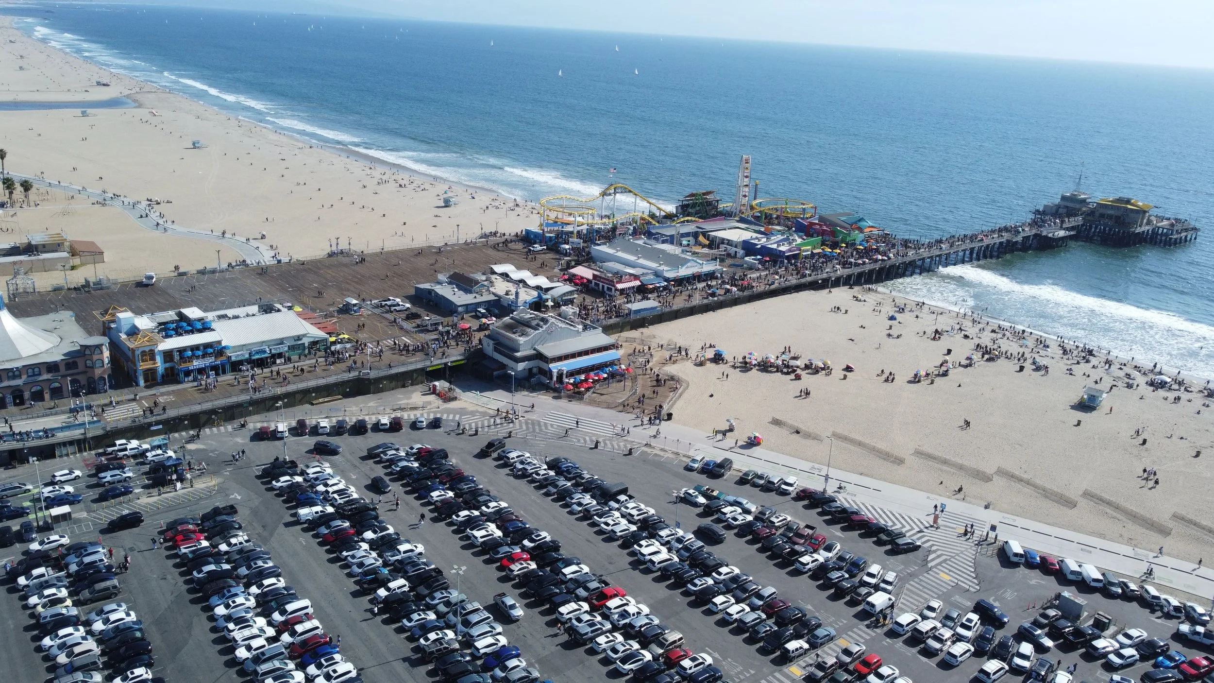 Aerial view of a busy beach and pier with parking lot in the foreground, sandy beach, amusement rides, and the ocean in the background.