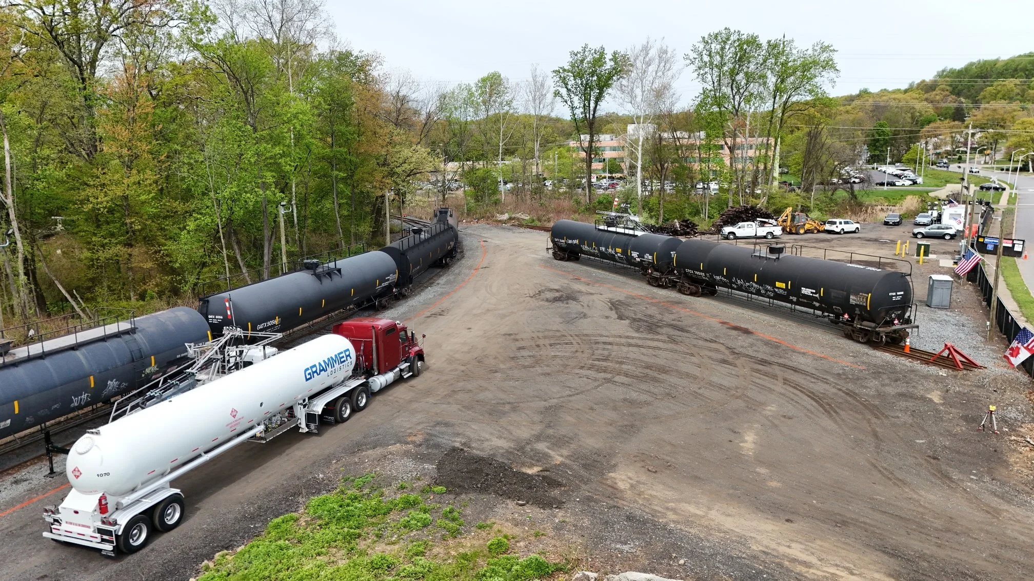 Aerial view of a train yard with several black tanker cars, a white fuel truck, and a parking lot in the background surrounded by trees and buildings.