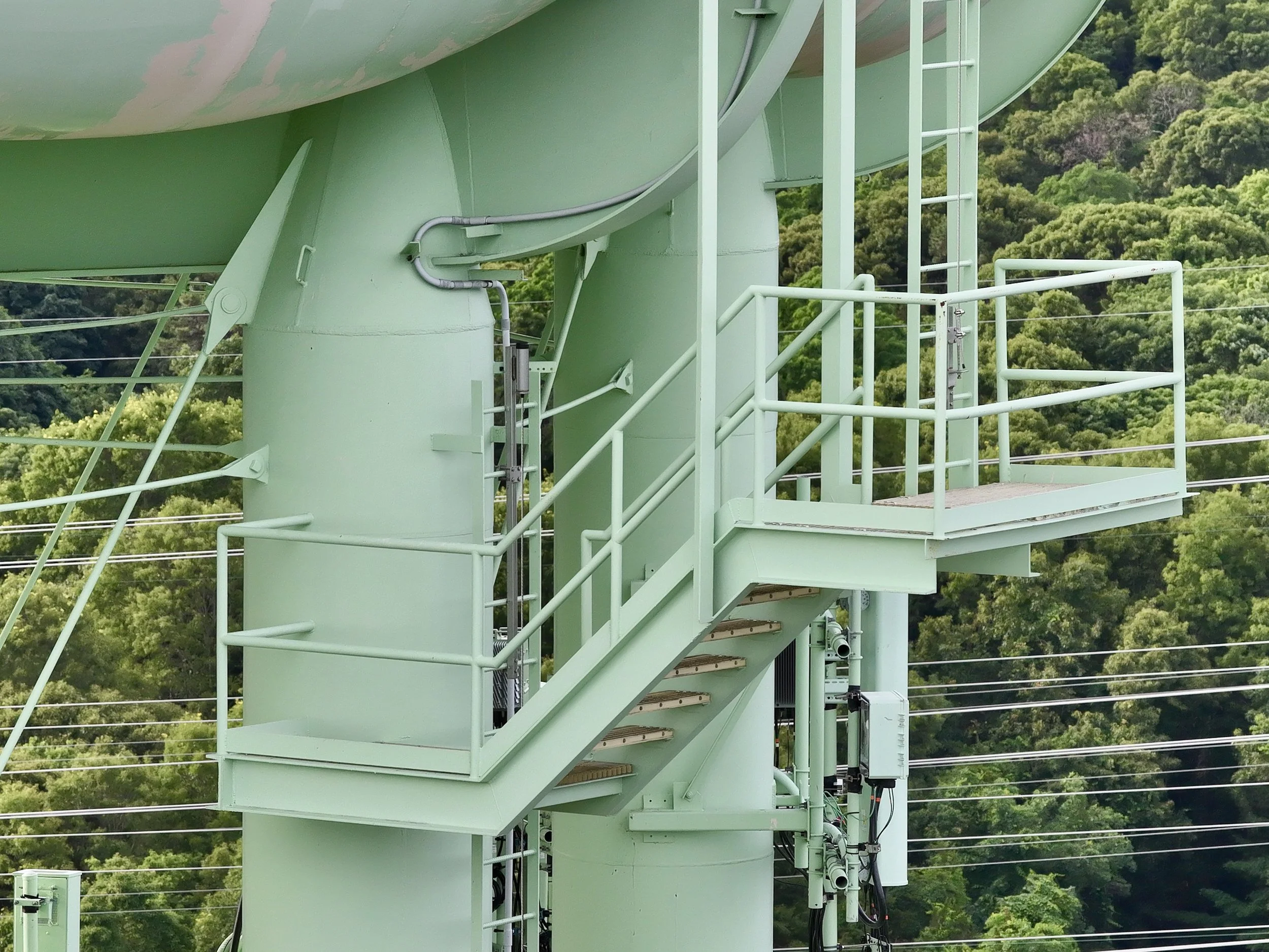 Close-up of a pale green metal structure with stairs and railings, part of a tower or observation platform, with trees and greenery in the background.
