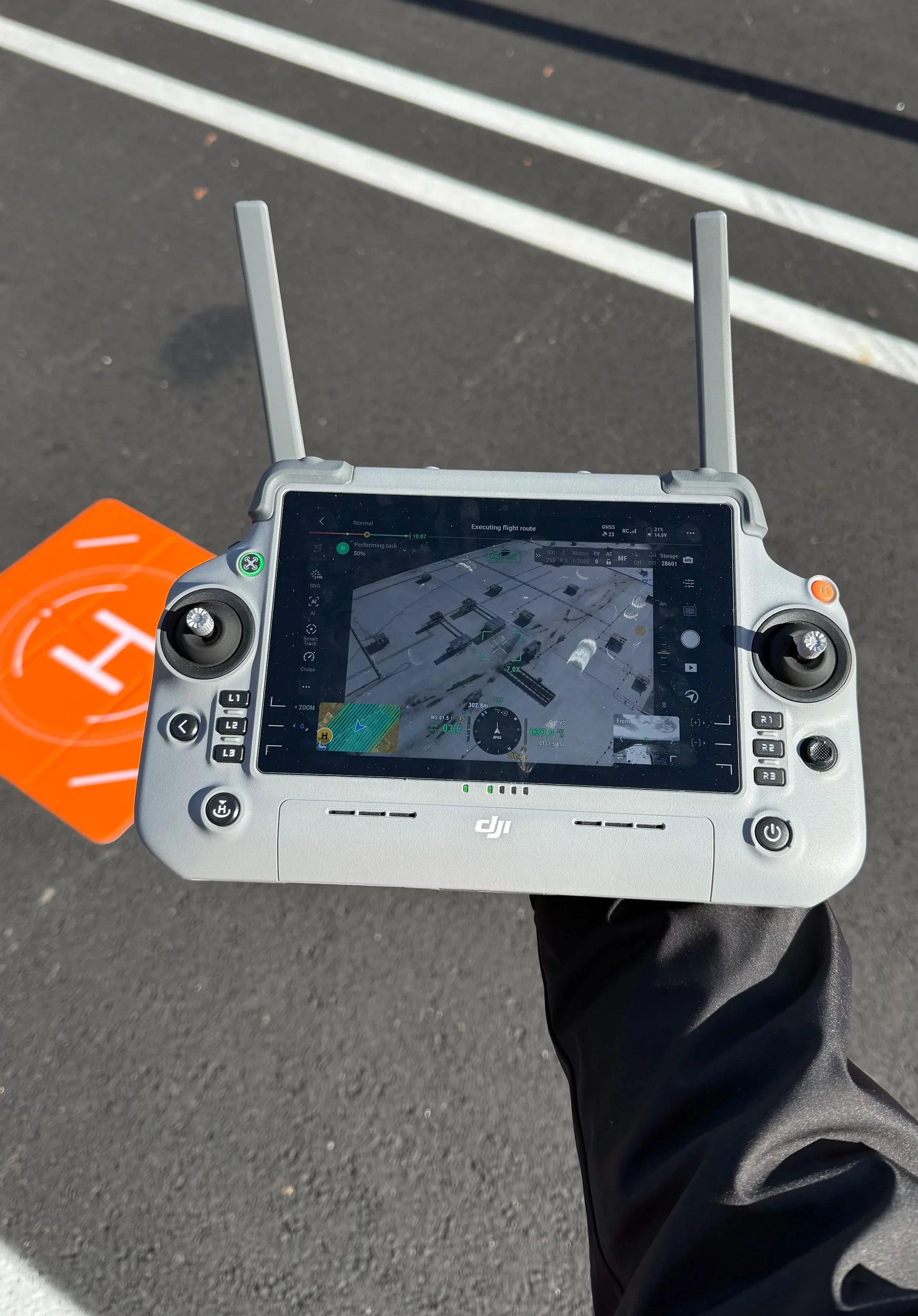 Drone remote controller with a tablet screen displaying a map, held by a person with a black sleeve in a parking lot with white lines and an orange drone landing pad.