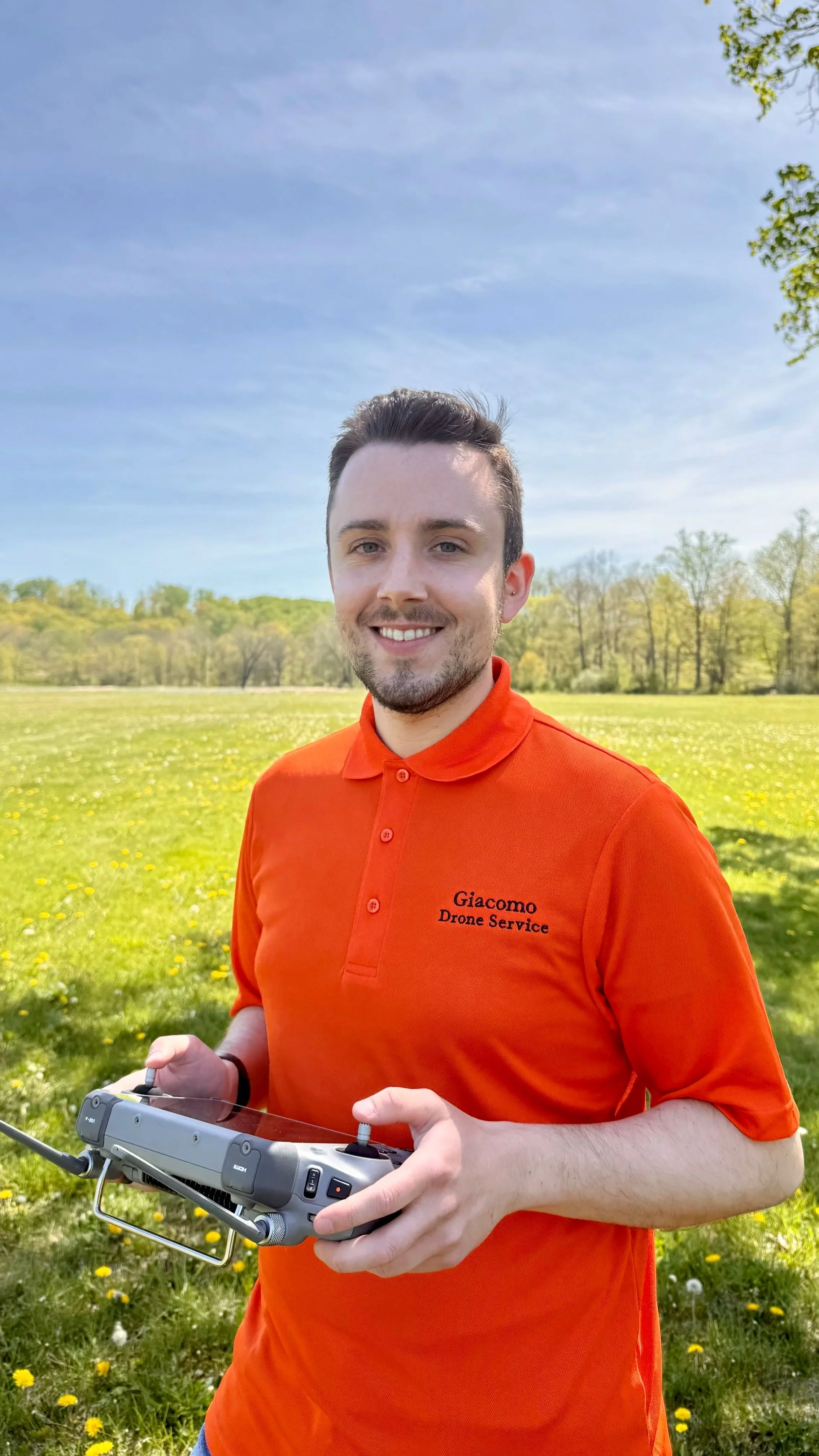 A young man with dark hair and a beard standing outdoors in a green field with trees in the background, wearing an orange polo shirt with 'Giacomo Drone Service' embroidered on it, holding a drone remote controller, smiling at the camera.