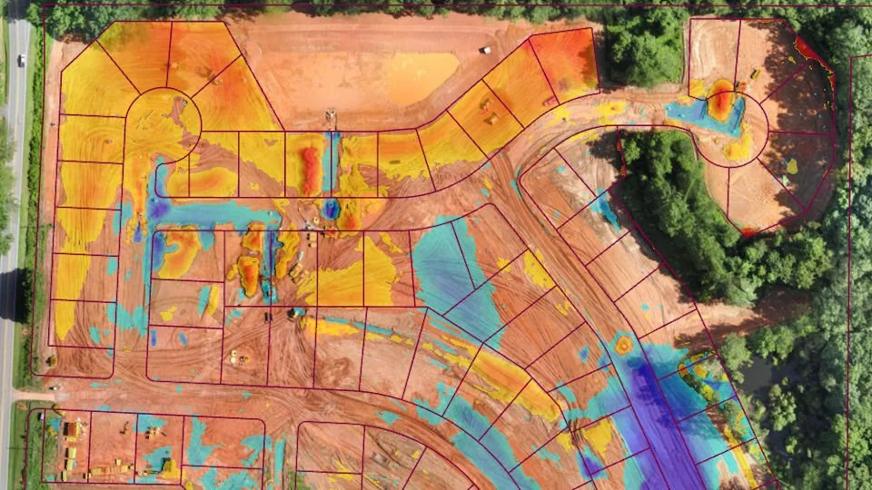 Aerial view of a residential construction site showing lots marked with colored markings indicating grading and drainage plans, with some areas covered in yellow and orange soil, and blue areas representing water flow or drainage features, surrounded by trees and partially developed infrastructure.