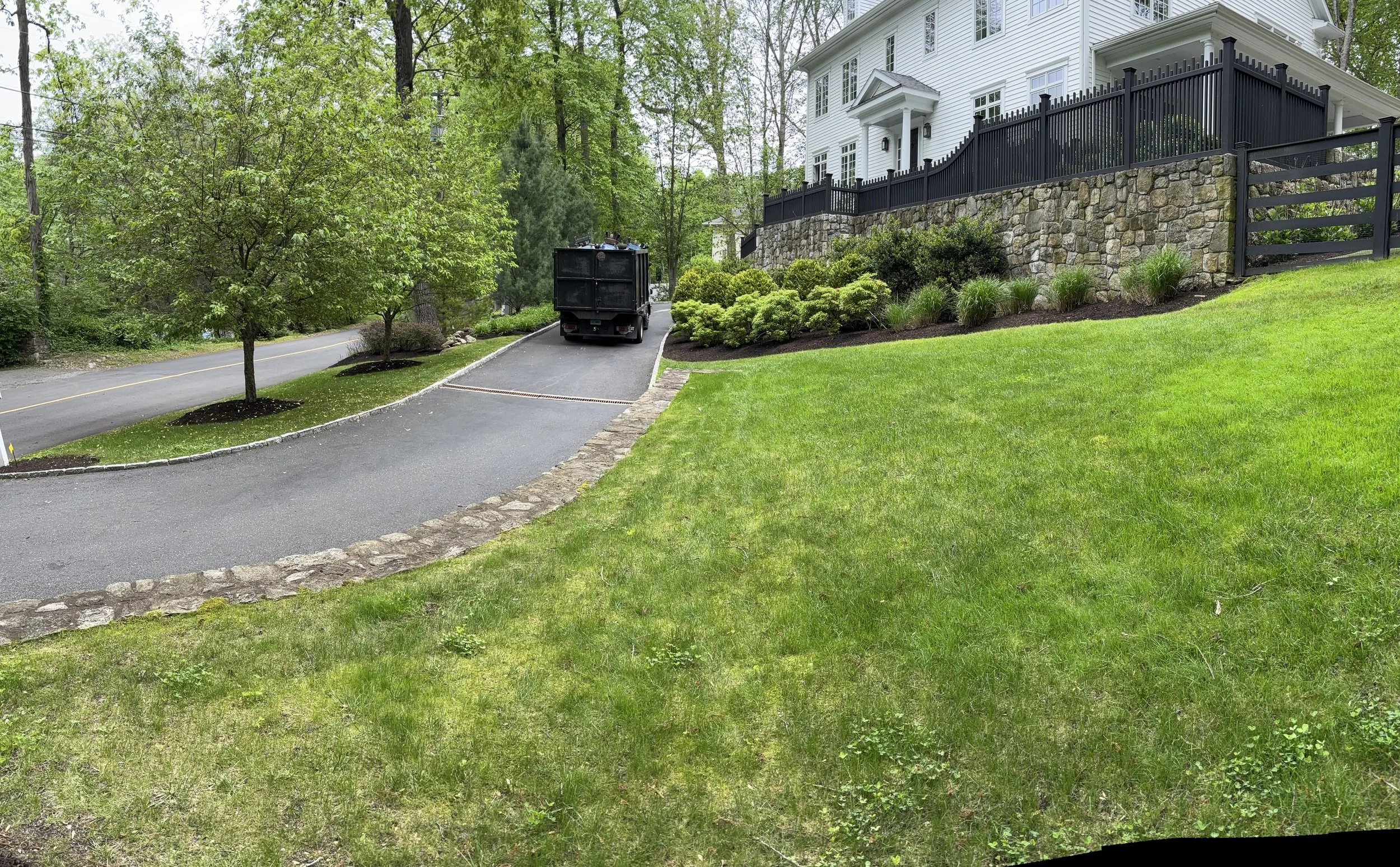 A residential yard with green grass, a stone border, and a paved driveway leading to a black truck. A white house with a black fence and stone wall is visible in the background, surrounded by trees.