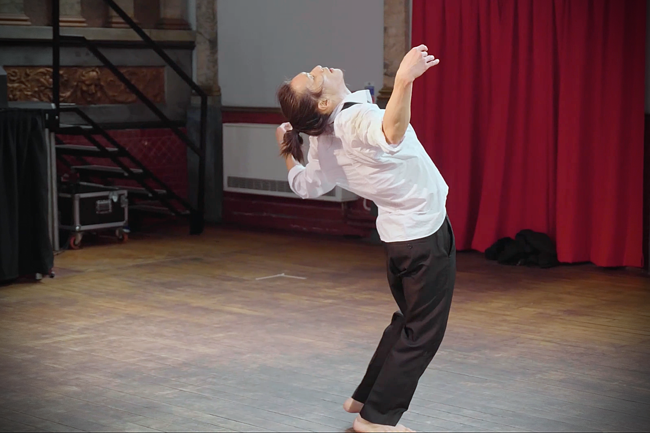 Dancer Kenny Ho dancing, mid-move, wearing a white shirt and black pants, on a wooden floor with red curtains in the background. From the dance theatre solo Innocence Lost.