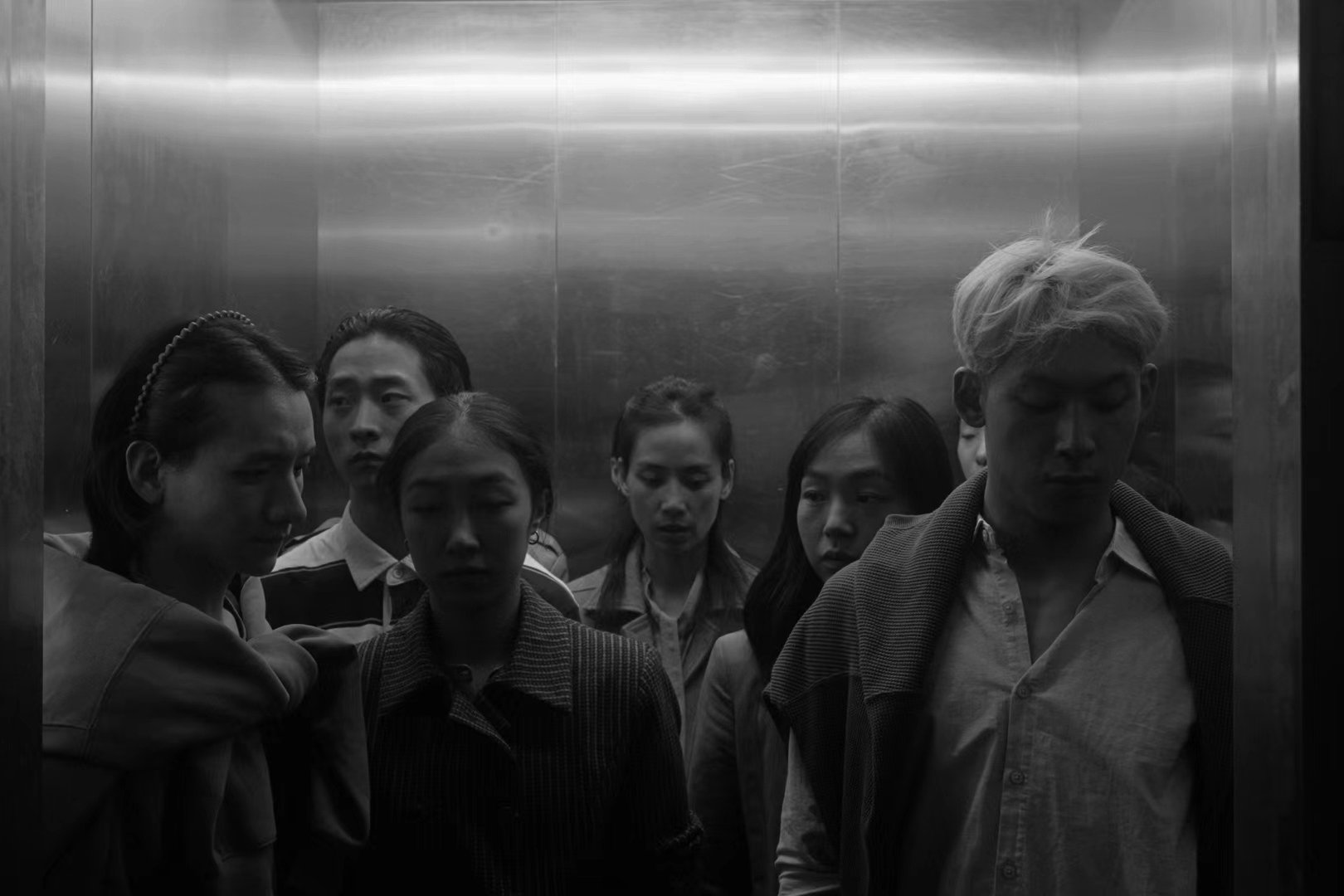 A group of six diverse people standing inside an elevator with metallic walls, looking downward or away, with serious or contemplative expressions.