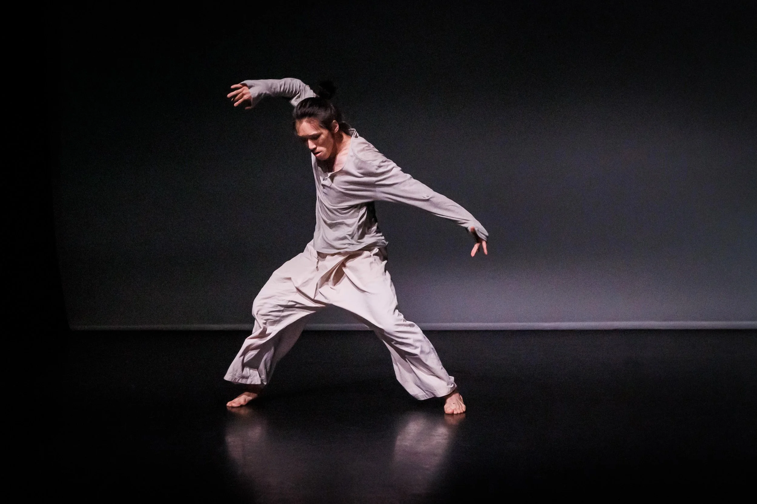 Kenny Wing Tao Ho performing a contemporary dance move on a dark stage, dressed in loose, light-colored clothing, with one arm raised and the other extended outward. From the dance solo ma.