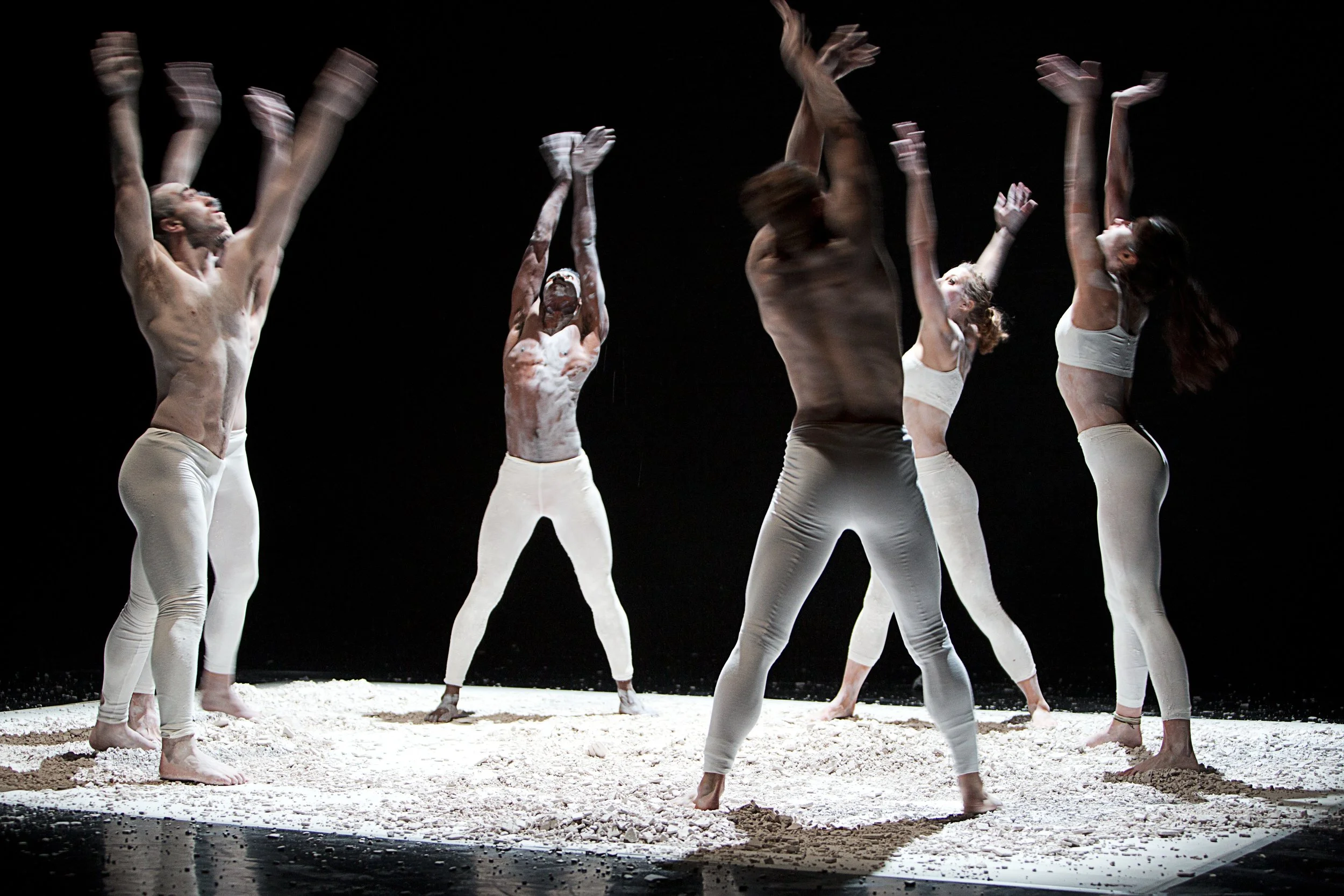 Six dancers with smeared white powder on their bodies, performing on a dark stage with white powder on the floor, dressed in white pants and tops, some with bare chests, striking expressive poses. From the choreography Dust to Dust.