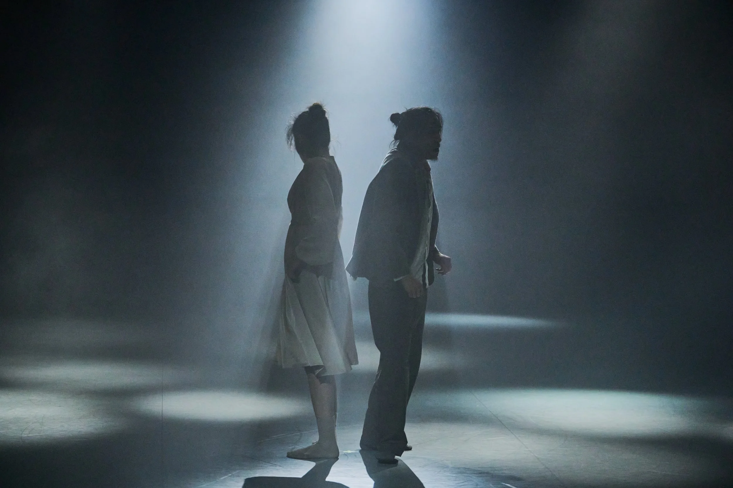 Dancers Yeji and Kenny silhouetted standing back to back in a dark, foggy space with a single light shining from above.