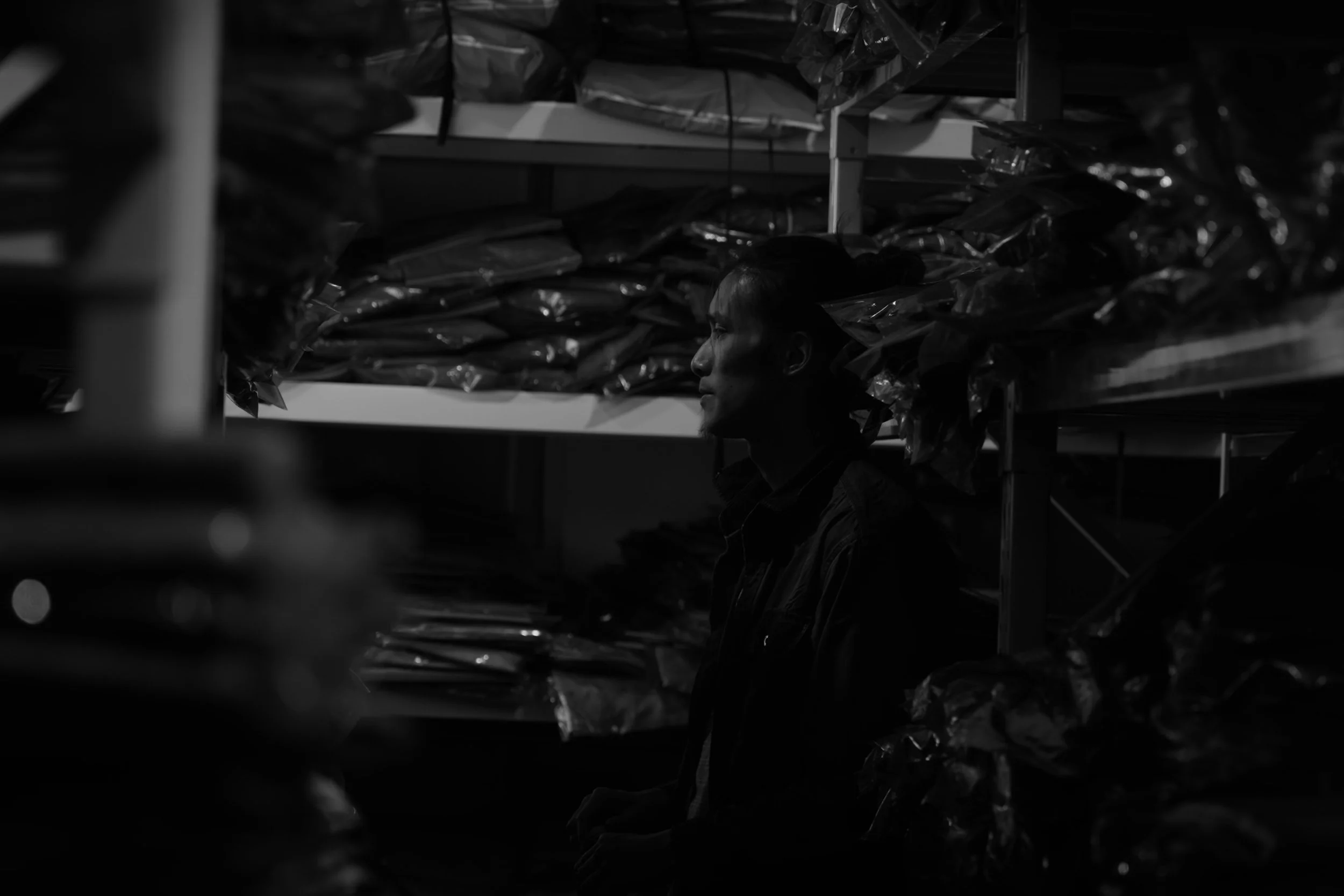 K leaning against a shelf in a warehouse, surrounded by bags of goods, in black and white. From the film Pigs in Heaven