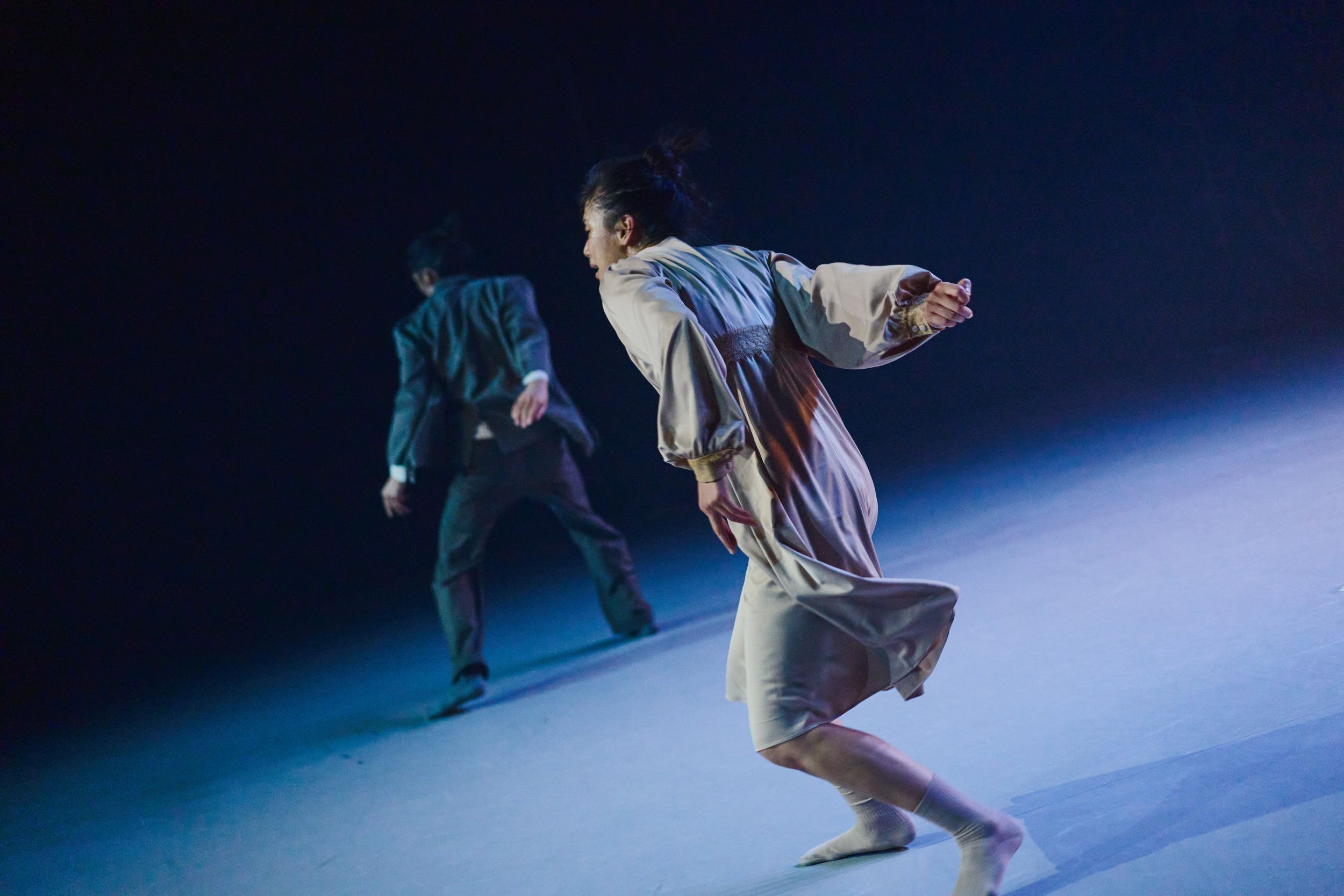 Dancers Yeji and Kenny performing on stage with dark background, Yeji in a satin dress and Kenny in a suit, captured in a contemporary dance work ARRIVAL