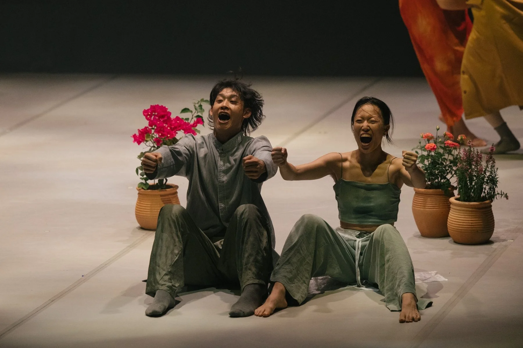 A man and a women sitting on the floor with engaged expressions, fists raised, surrounded by potted plants, in a dimly lit setting. From the dance theatre production Flowers of Ruin.
