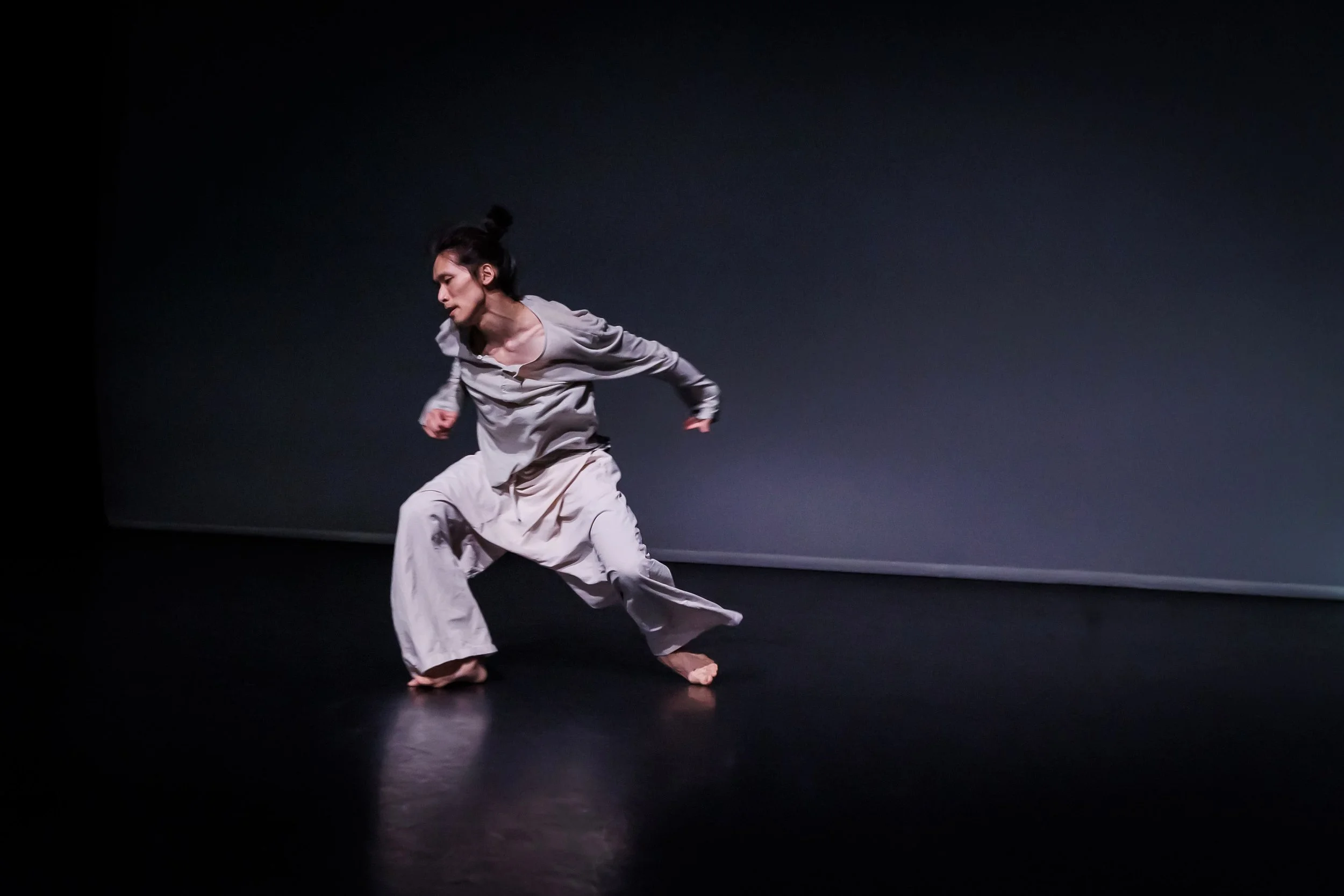 Dancer Kenny Ho is performing on stage in a gray outfit, with a dark background and minimal lighting. From the dance solo ma.