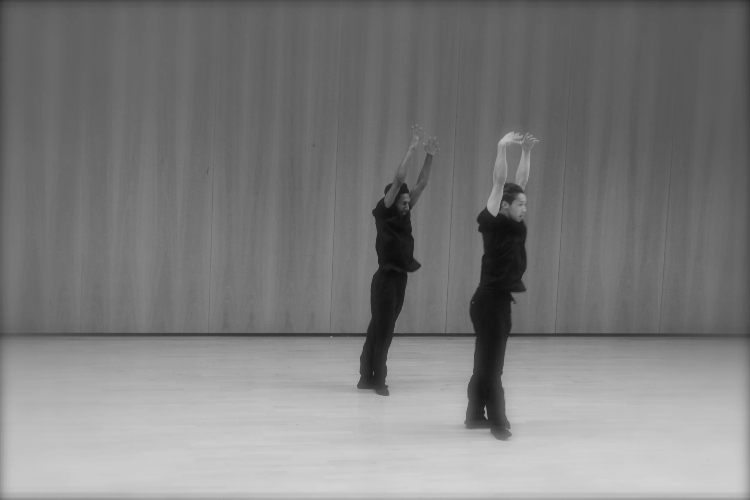 Dancers Kenny and Ihsaan in black clothing dancing in a studio with a wooden wall background. From the choreography Casting Shadows.