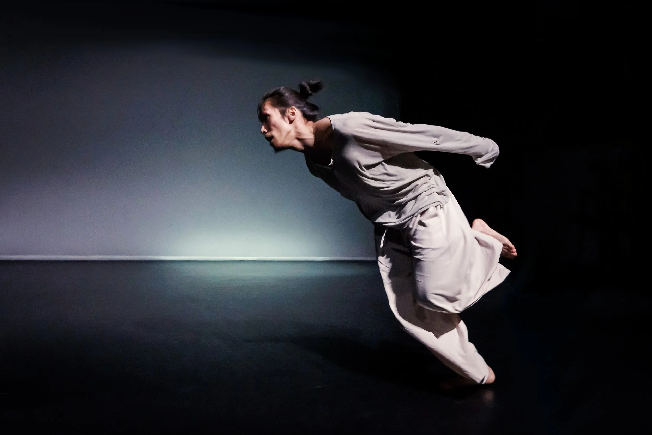 Kenny Wing Tao Ho dressed in light-colored clothing is in a dance on stage, leaning forward and runningwith his arms behind his back. From the dance solo ma.