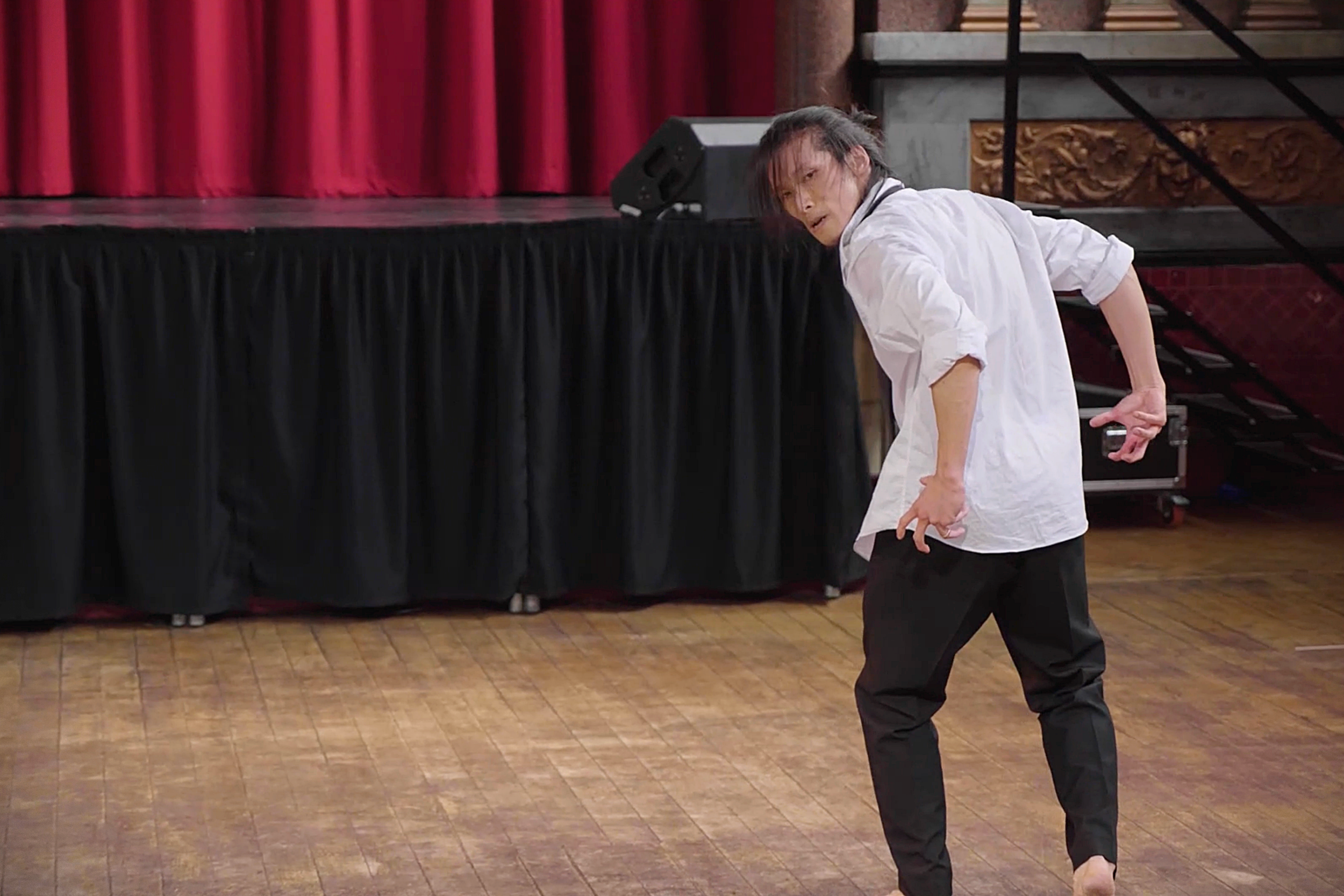 Kenny Ho in white shirt and black pants dancing on wooden floor in front of stage with red curtain. From the dance theatre solo Innocence Lost.
