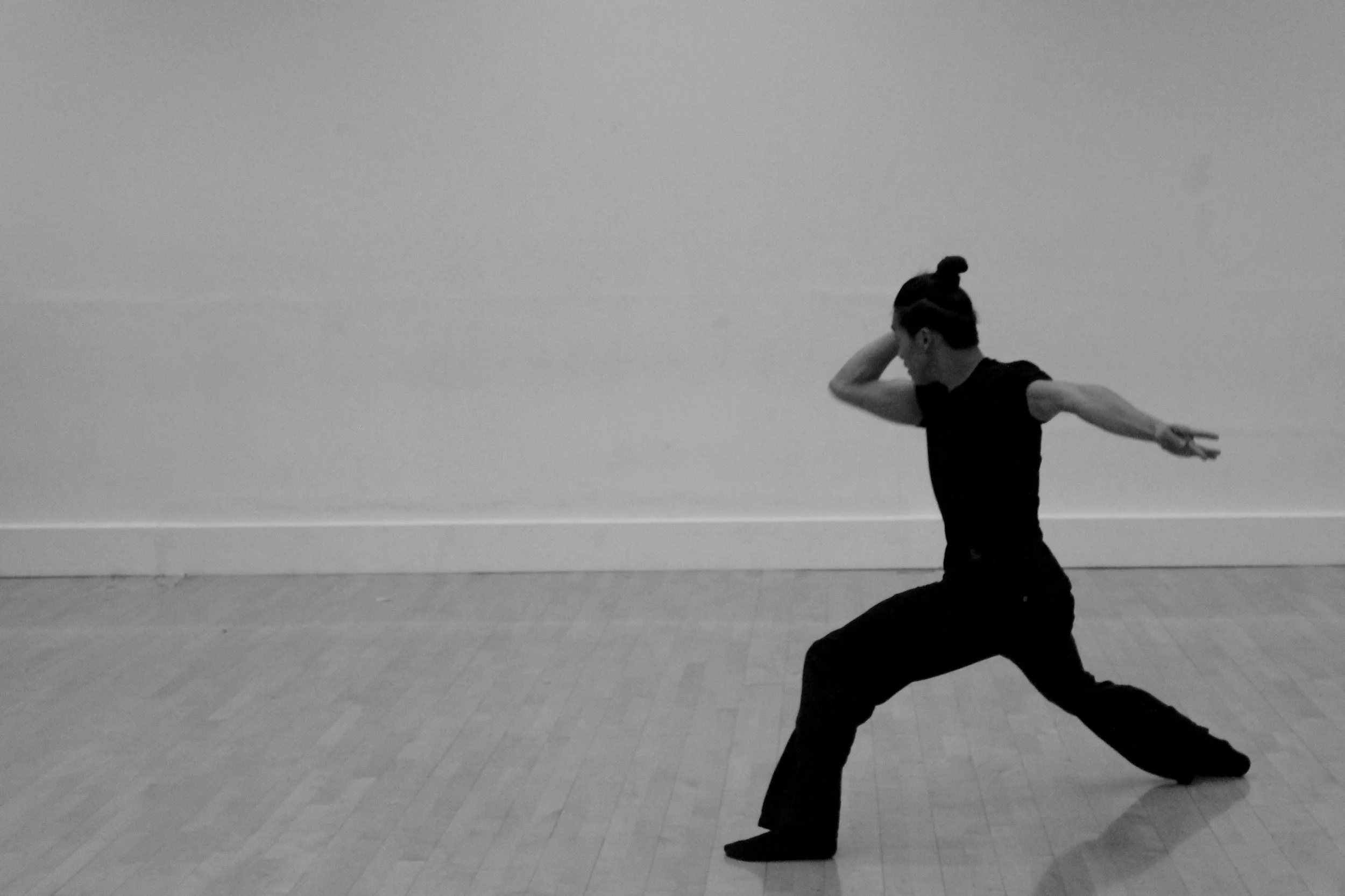 Kenny Ho dancing in a studio, wearing black clothing, against a plain wall. From the choreography Casting Shadows.