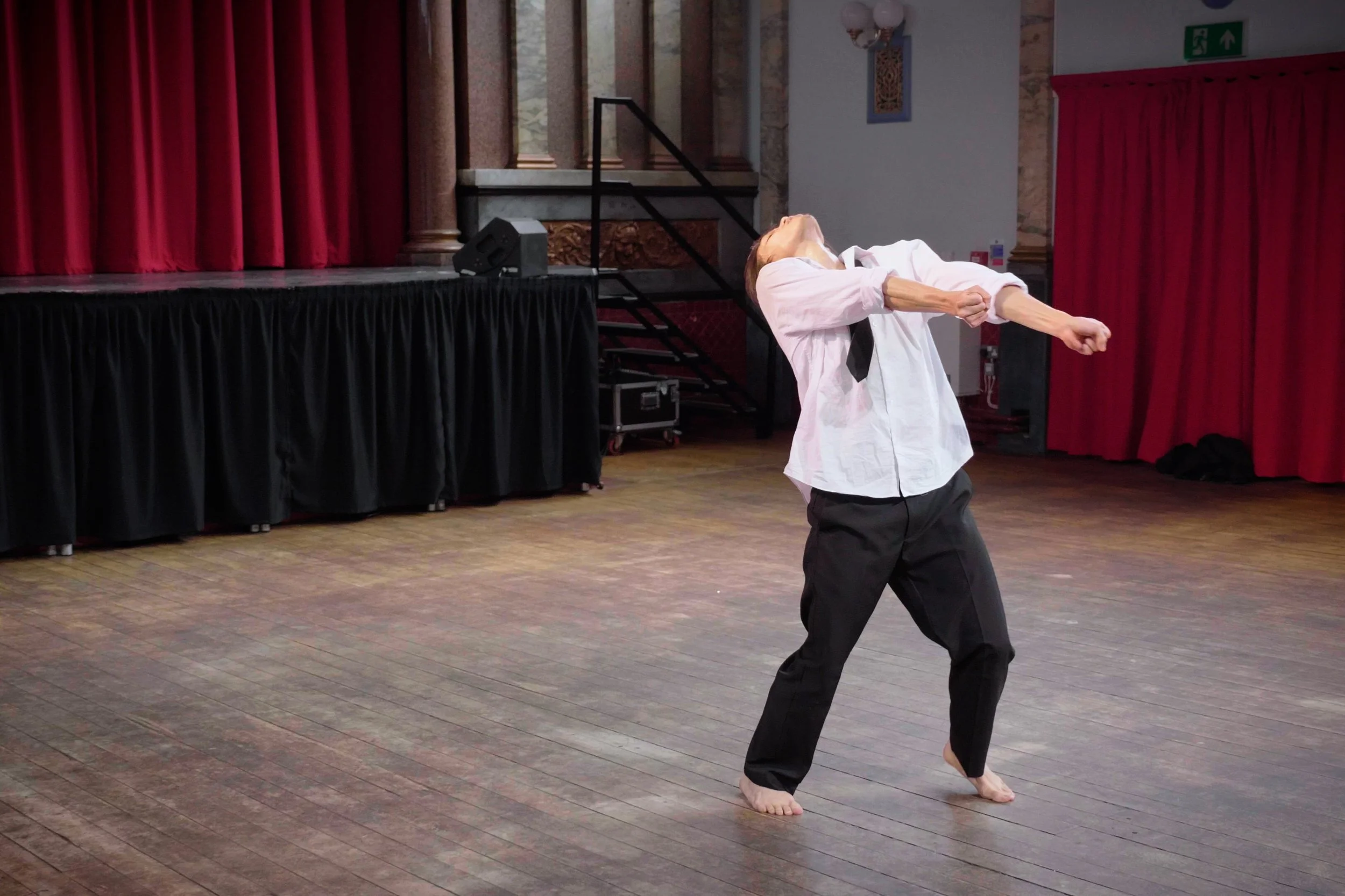 Dancer Kenny Ho performing a dance move on a wooden floor with red curtains and a stage in the background. From the dance theatre solo Innocence Lost.