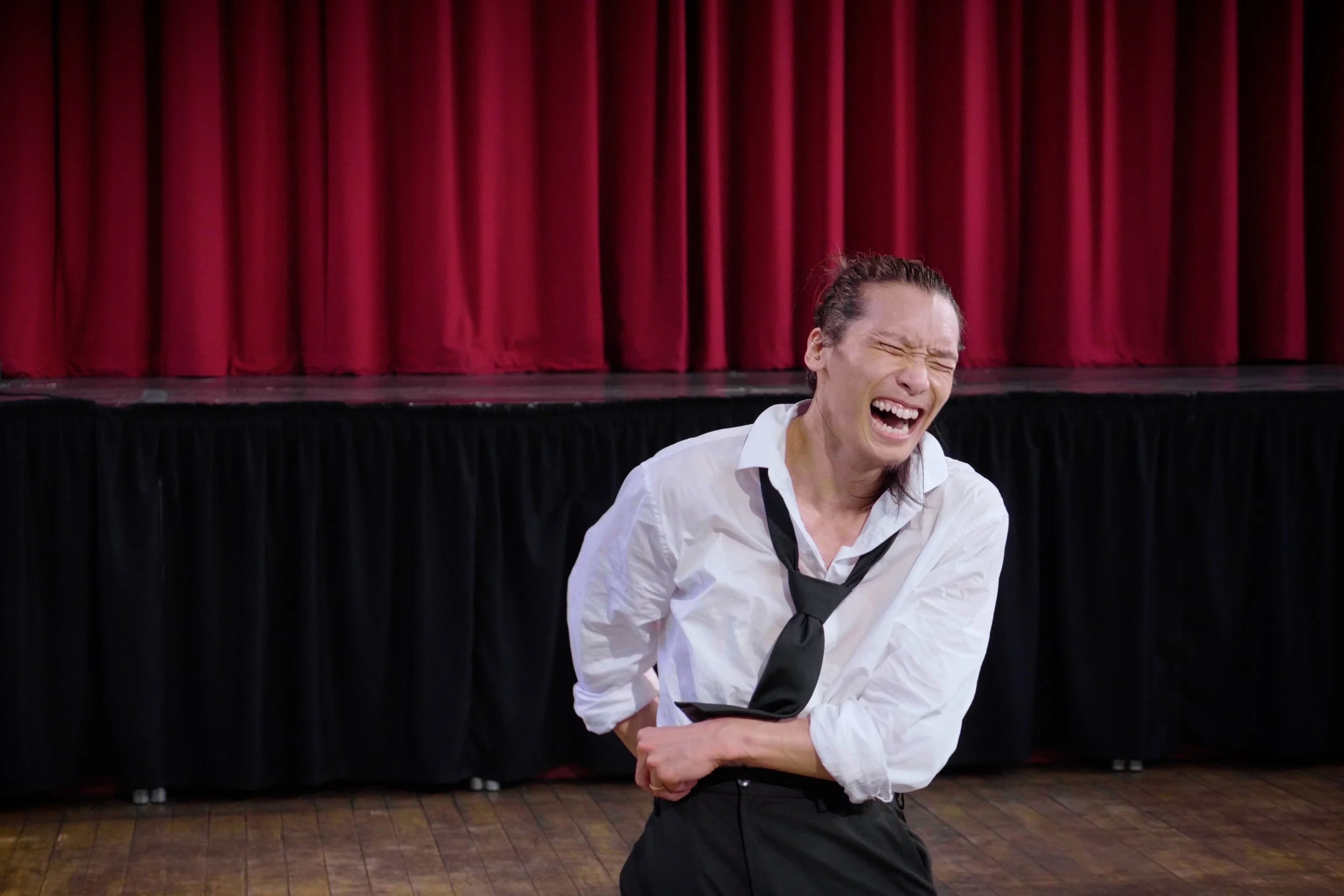 Dancer Kenny Ho in a white shirt and black tie, standing in front of red curtains and a black stage skirt, with eyes closed and mouth open laughing. From the dance theatre solo Innocence Lost.