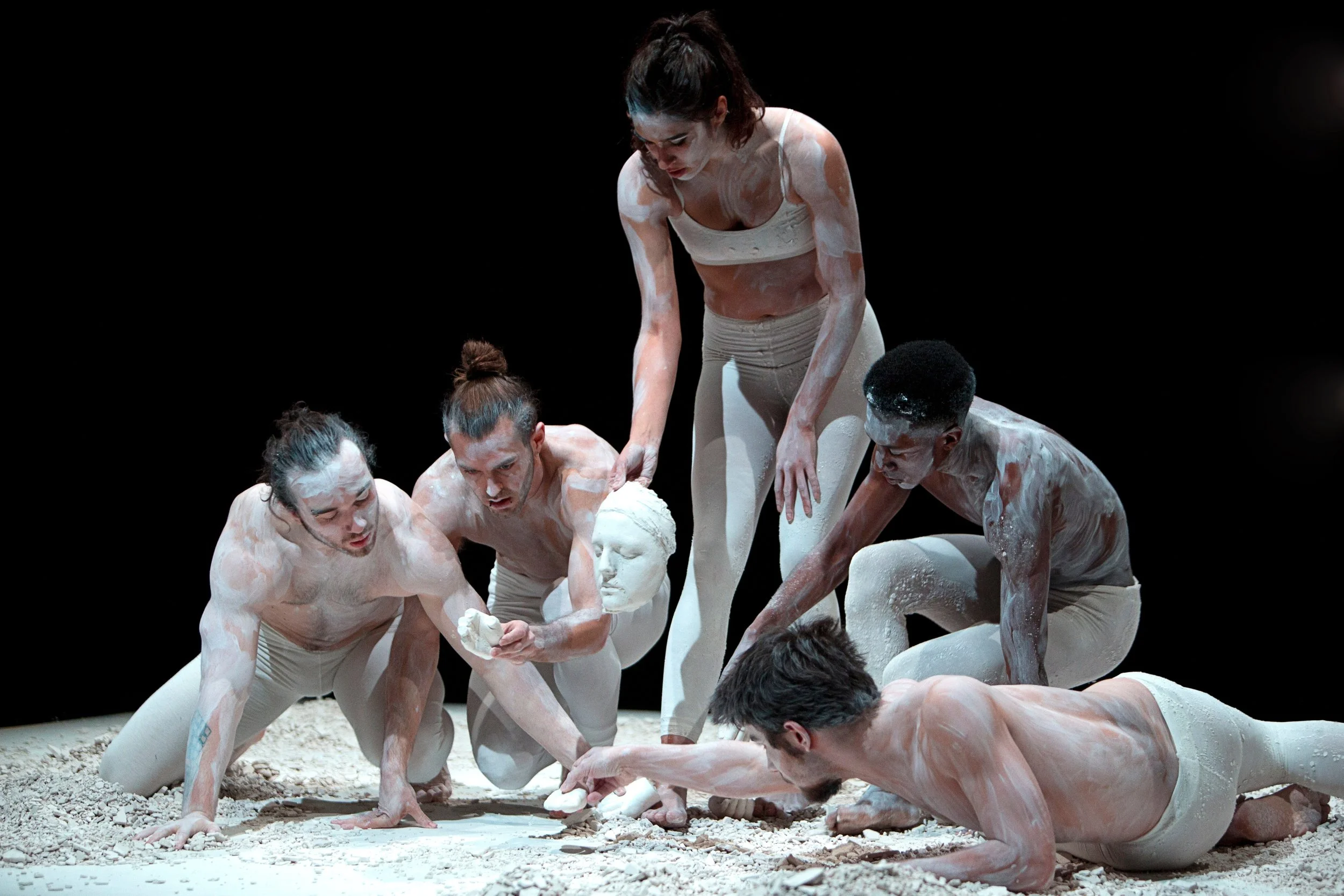 Group of five performers covered in white powder, engaged in a contemporary dance each holding a fragmented white sculpture on a dark stage. From the choreography Dust to Dust.