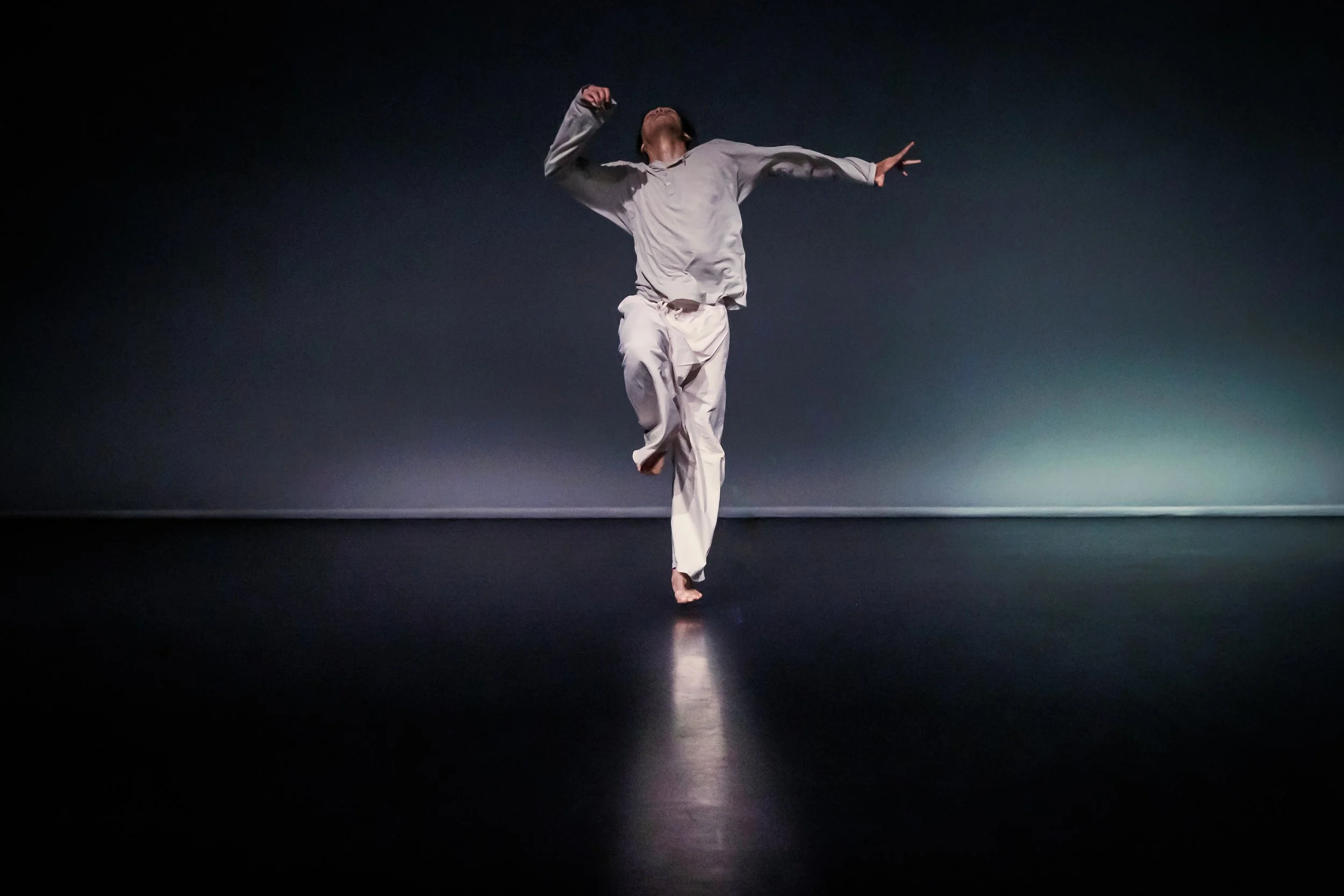 Kenny Wing Tao Ho dancing on a dark stage, wearing loose light-colored clothing, jumping with one foot off the ground and arms extended up and to the side. From the dance solo ma.