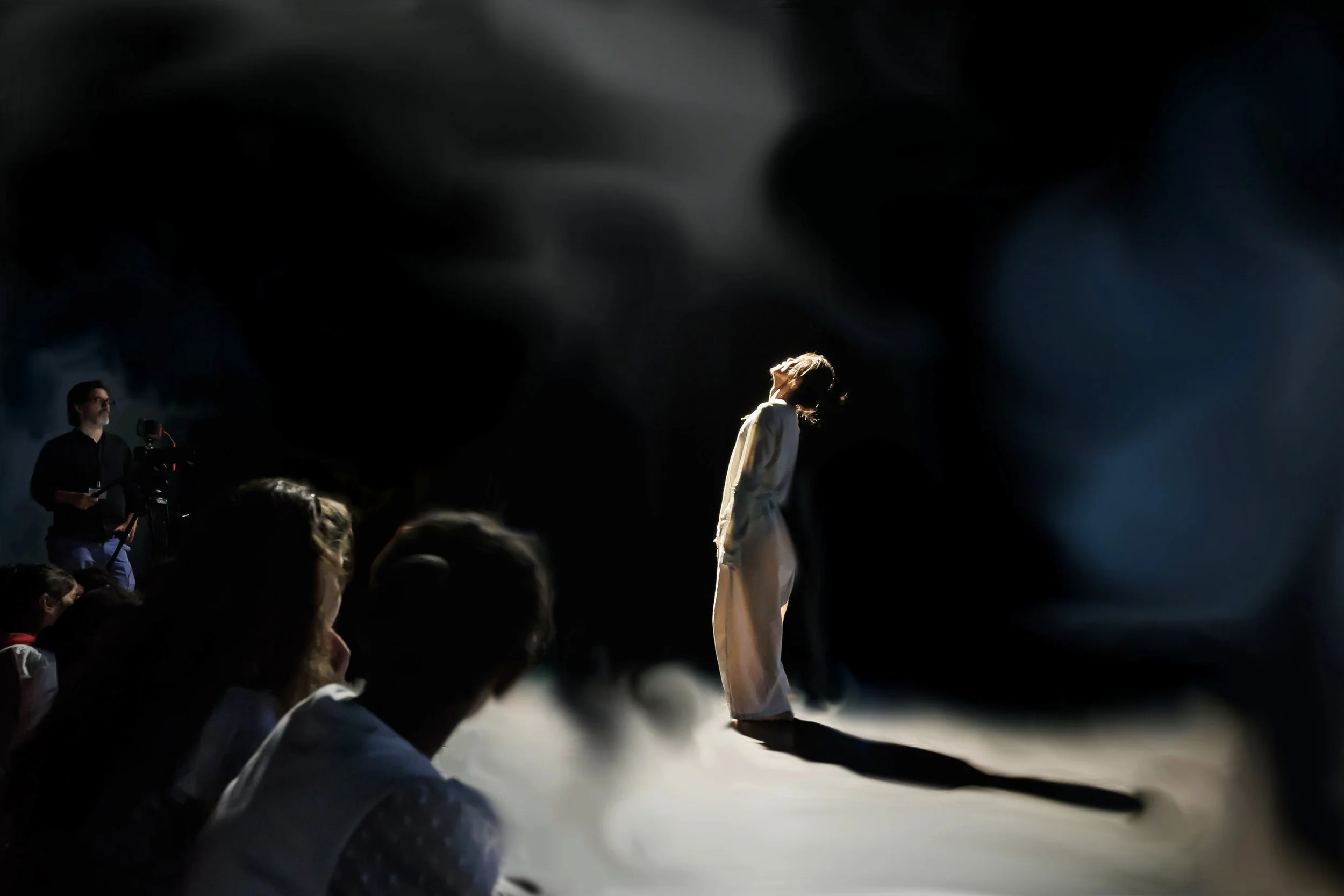 Kenny Wing Tao Ho in a beige outfit standing on a dark stage, illuminated by a spotlight, with an audience observing him. From the dance solo ma.