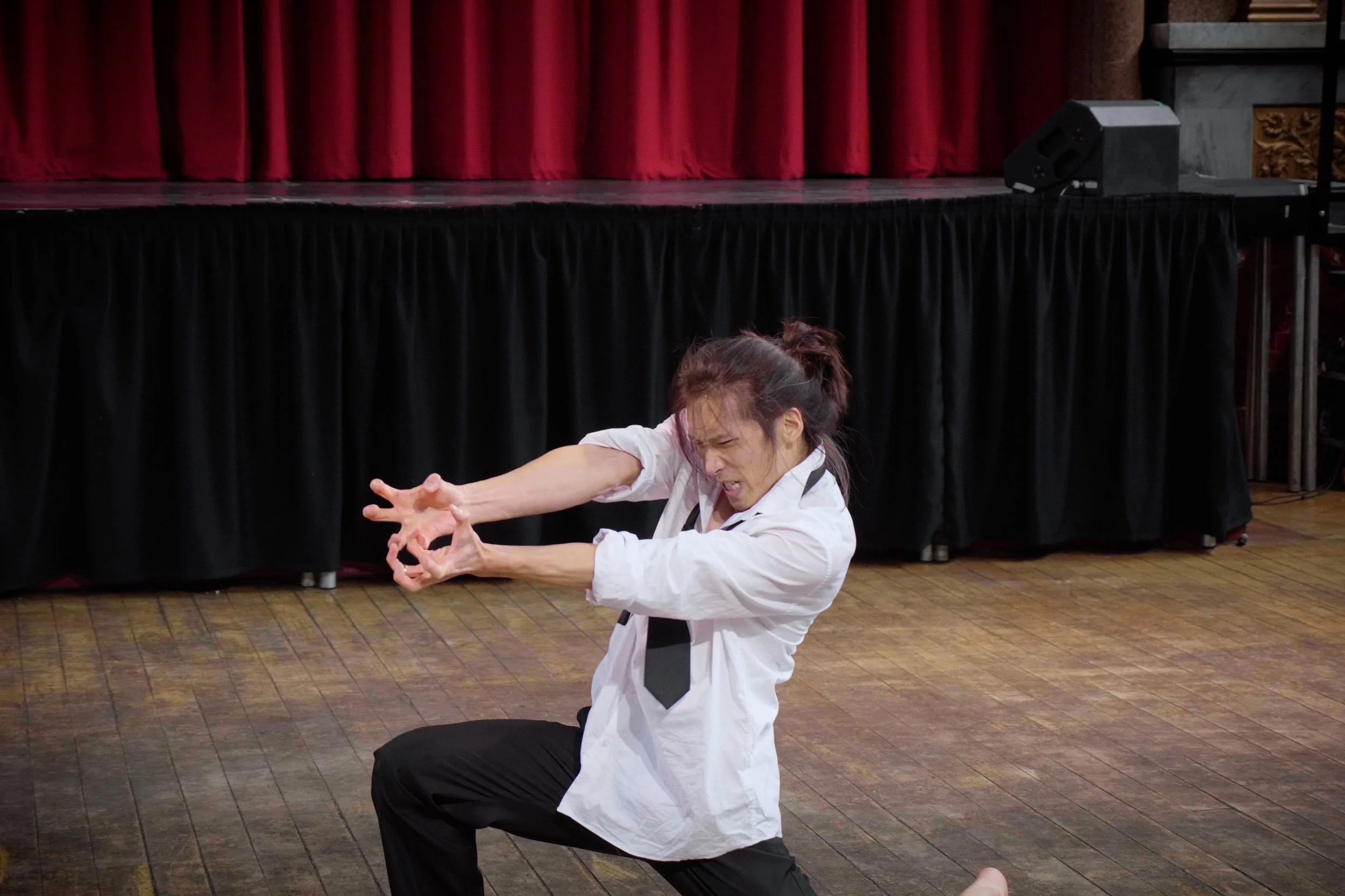 Kenny Ho performering in a white shirt and black pants on stage, making a dramatic gesture with her arms and hands. From the dance theatre solo Innocence Lost.
