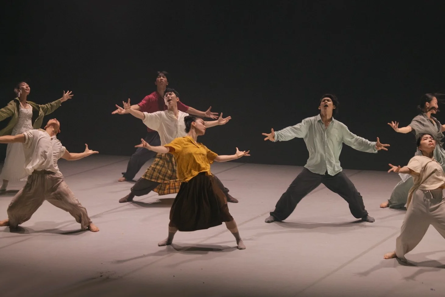 Group of dancers performing on stage with arms outstretched, wearing pastel and bright coloured clothing. From the dance theatre production Flowers of Ruin.