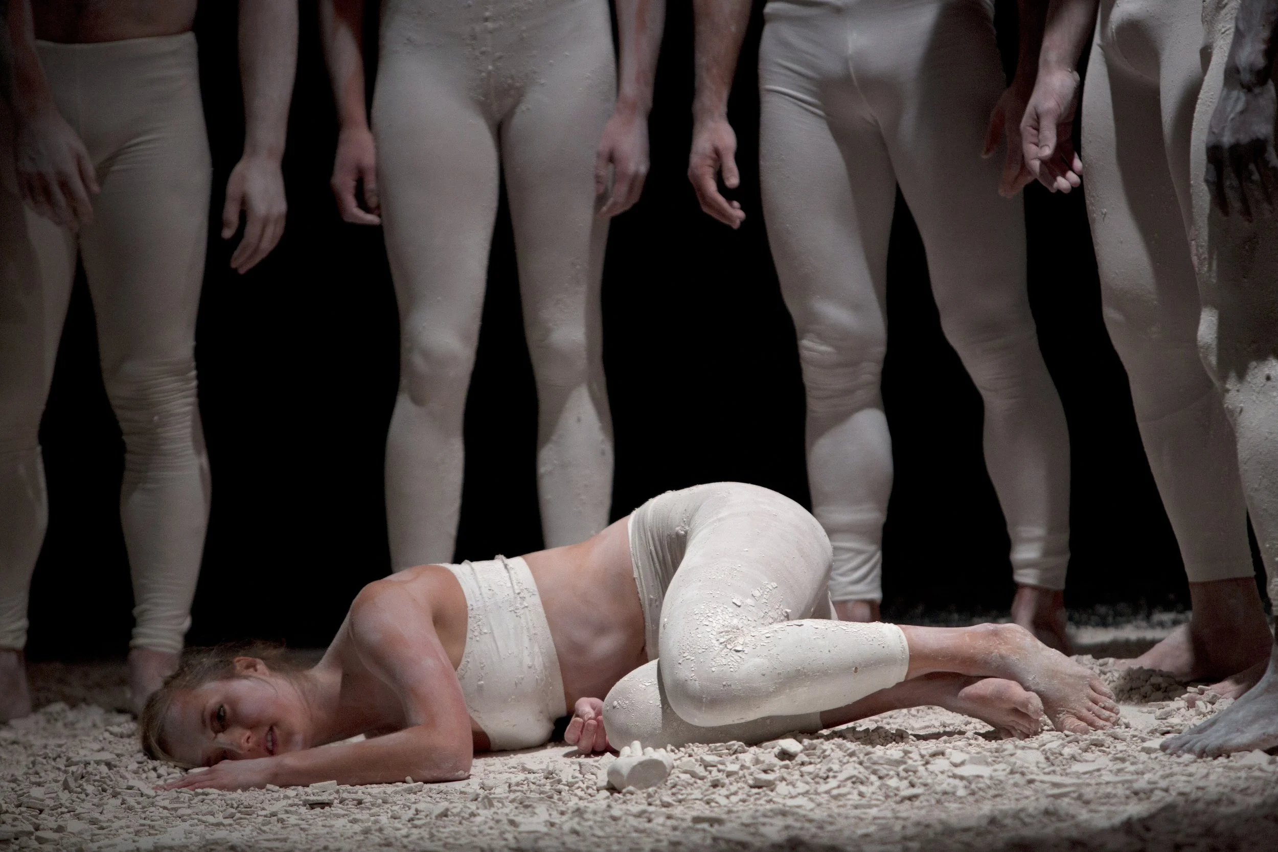 A woman lying on the ground covered in white clay fragments, surrounded by standing people in white leggings and tops, with a dark background. From the choreography Dust to Dust.