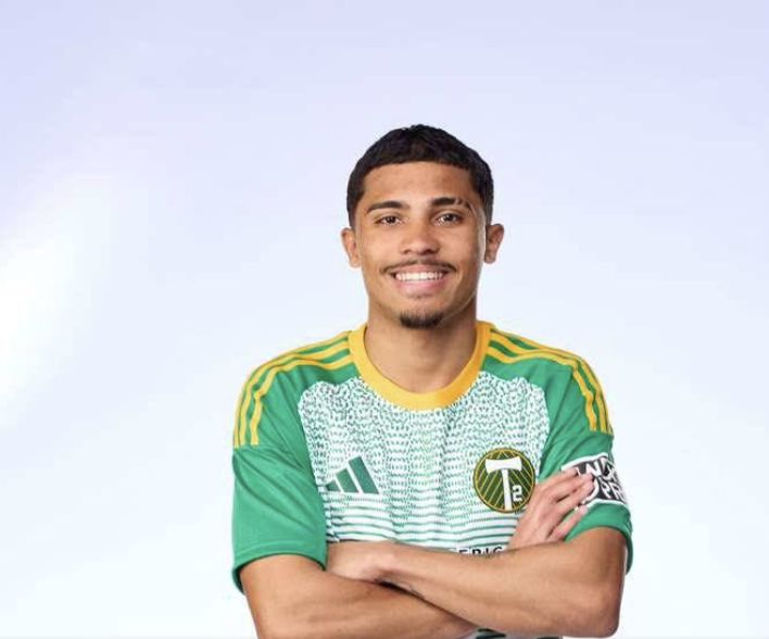 A man with short black hair and a goatee, smiling, wearing a green and yellow sports jersey with a team logo, standing with arms crossed against a light blue background.