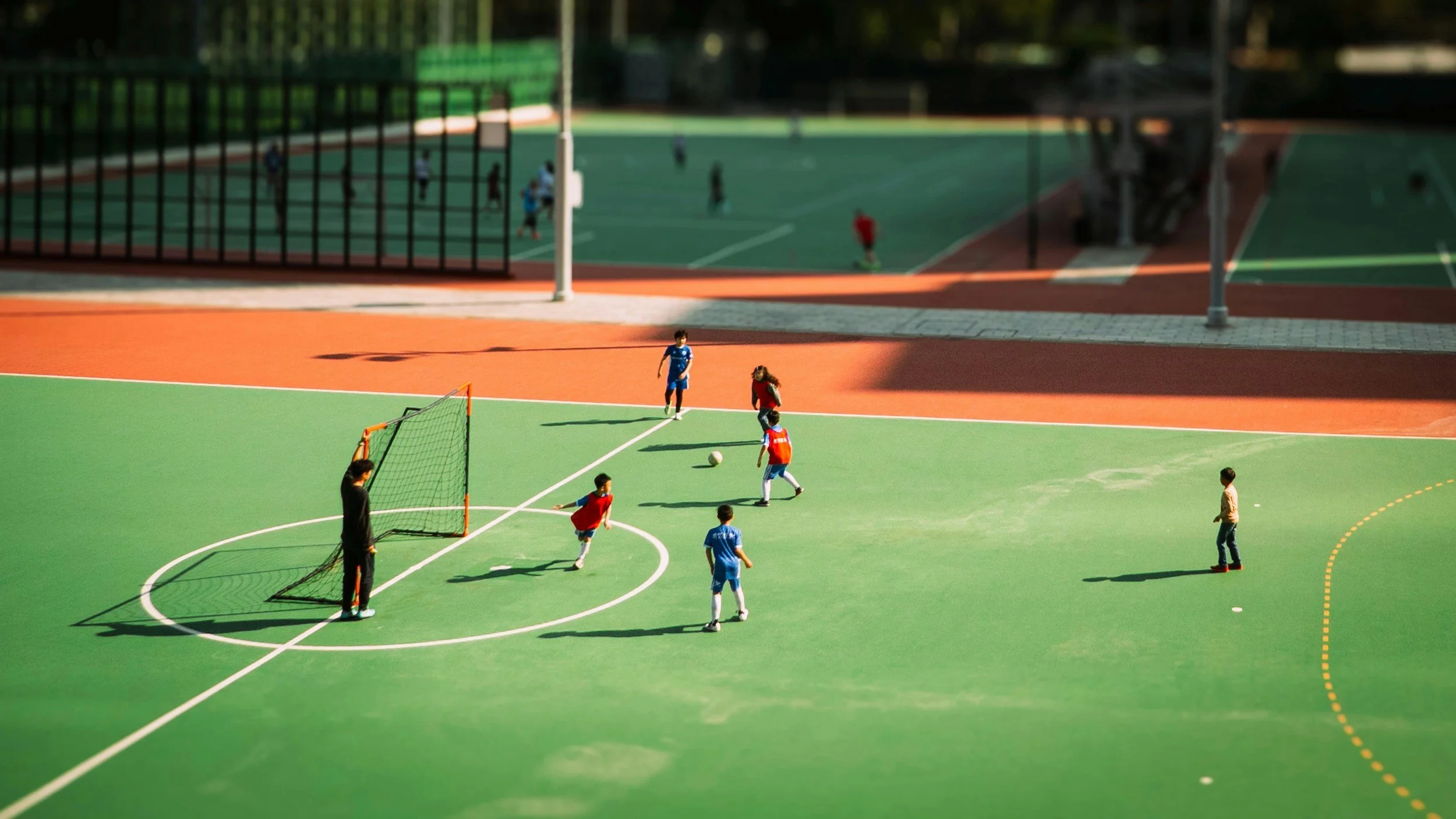 Children playing soccer on a green outdoor court, with a small goal, surrounded by a red track and other sports courts in the background.