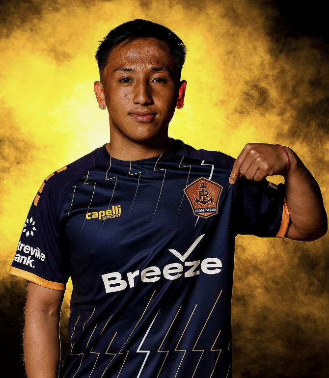 A young man with short black hair, earrings, and a red bracelet on his wrist, wearing a dark blue sports jersey with orange accents, holding up the Rhode Island FC team logo on his chest. The background is a yellow and black smoky pattern.