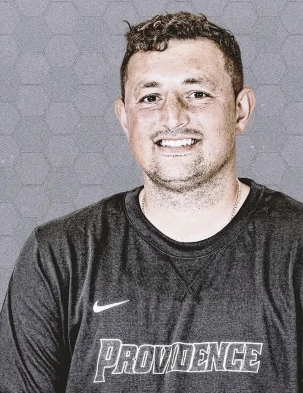 A young man sitting on a bench against a beige wall, wearing a gray polo shirt and cream-colored pants, with a serious expression and shadow cast on the wall.