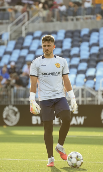 Soccer goalkeeper standing on the field with a soccer ball at his feet, wearing a white jersey with the logo of a sports team, dark shorts, and gloves, in a stadium with blue and black seats.