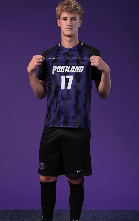 Young male soccer player wearing a Portland basketball jersey with number 17, black shorts, and black knee-high socks, standing against a purple background.