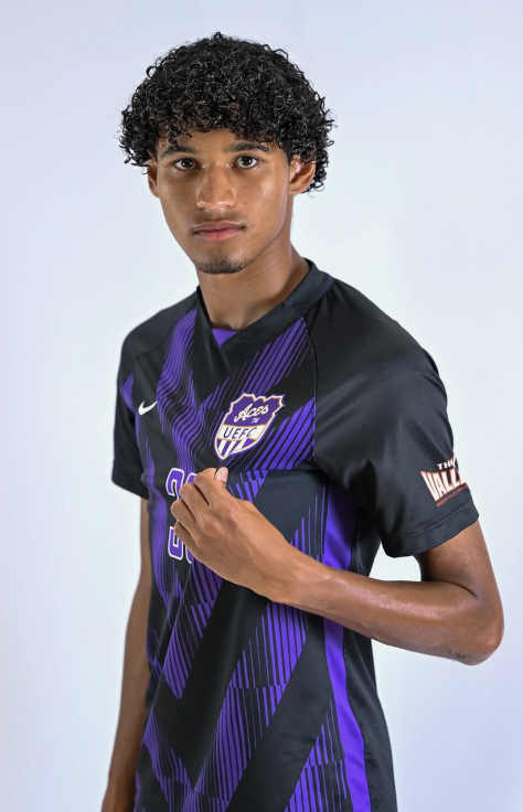 Young man in a black and purple soccer jersey with logos, standing against a light background.