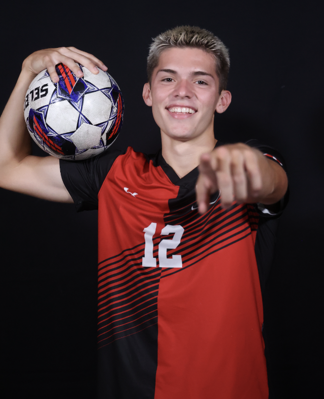 Young male soccer player holding a soccer ball over his shoulder, pointing at the camera, wearing a red and black jersey with the number 12, on a black background.