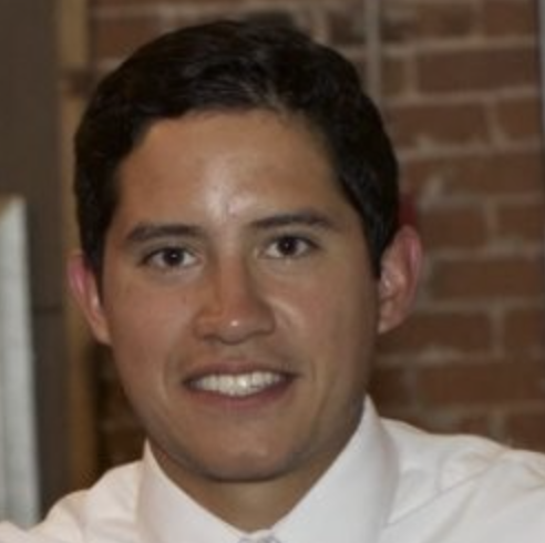 A young man with dark hair, wearing a white shirt, smiling, in front of a brick wall.